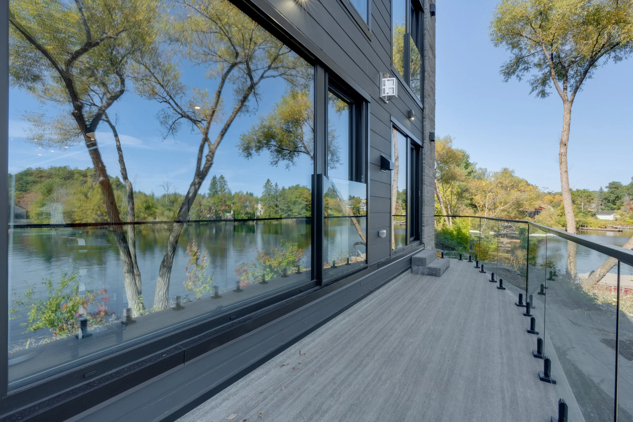 Balcony with glass railing overlooking a lake and trees with green and yellow leaves.