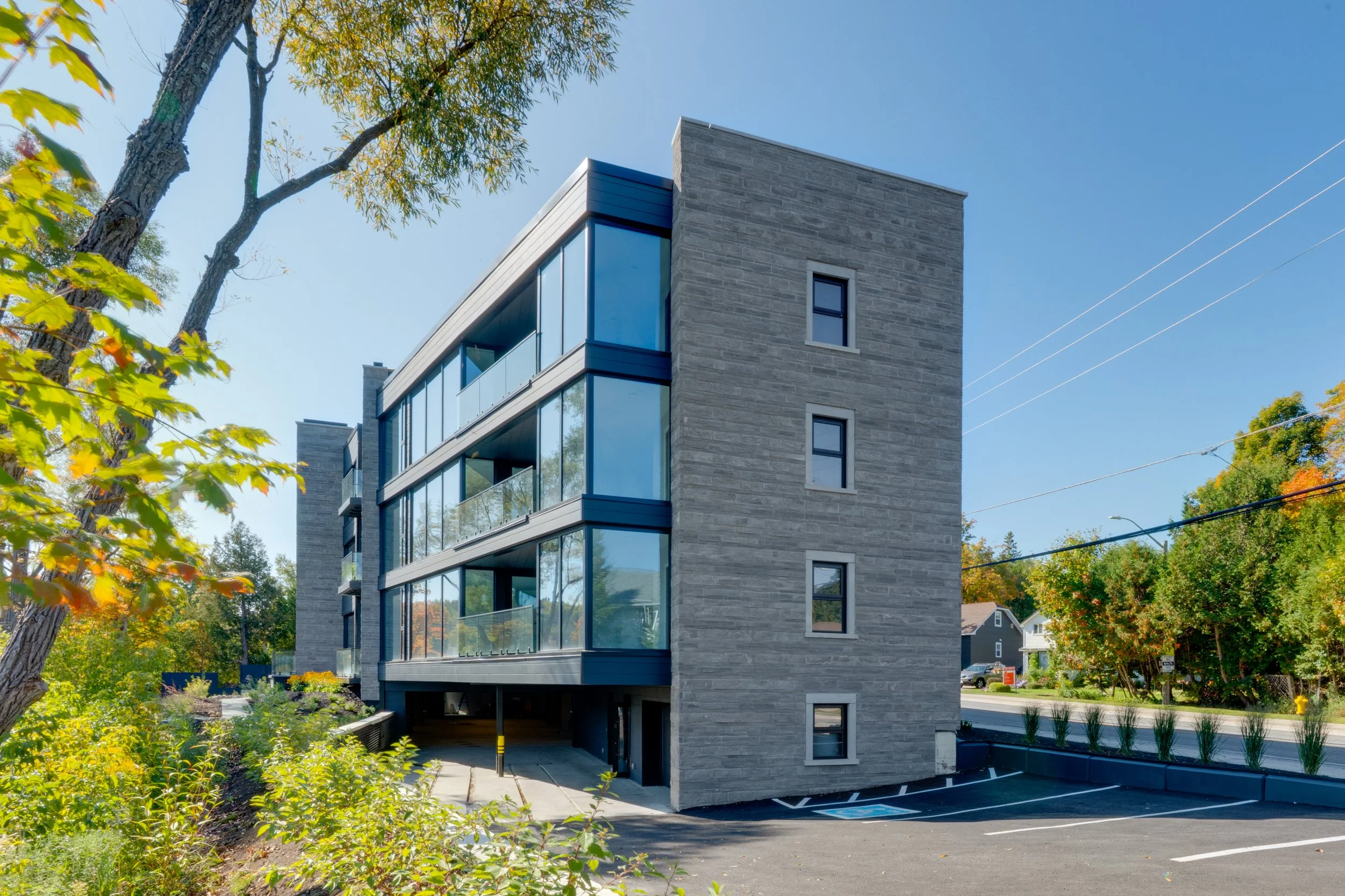 Modern multi-story apartment building with large glass windows and gray brick exterior, surrounded by trees and a parking lot.