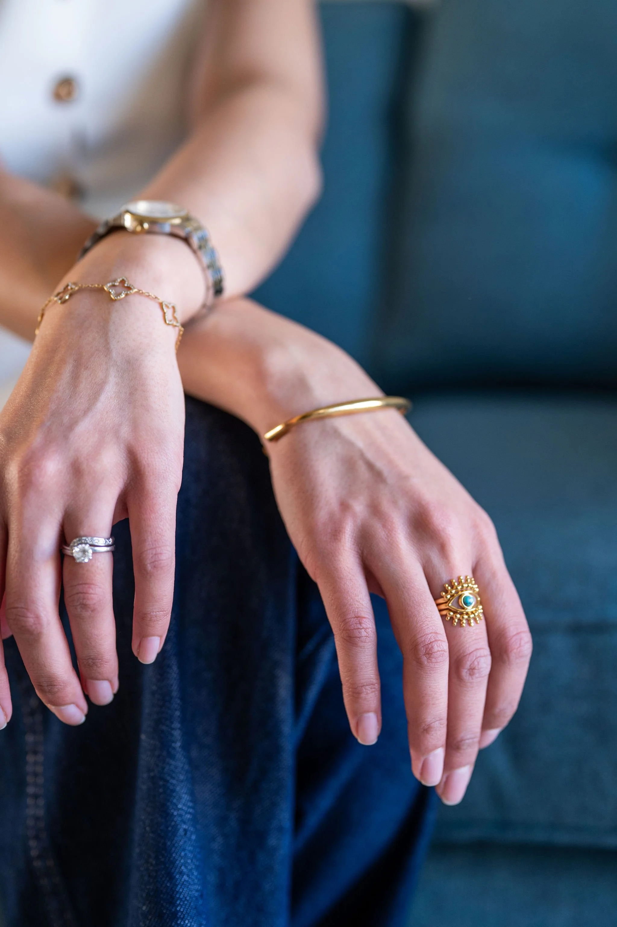 Close-up of a person's hands adorned with various rings and bracelets, resting on their lap with a blue couch in the background.