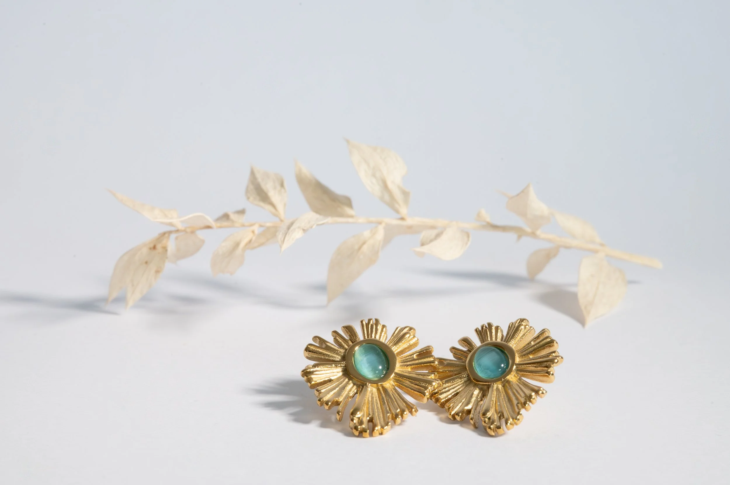 Pair of gold earrings with green gemstone centers on a white surface, with dried white leaves in the background.