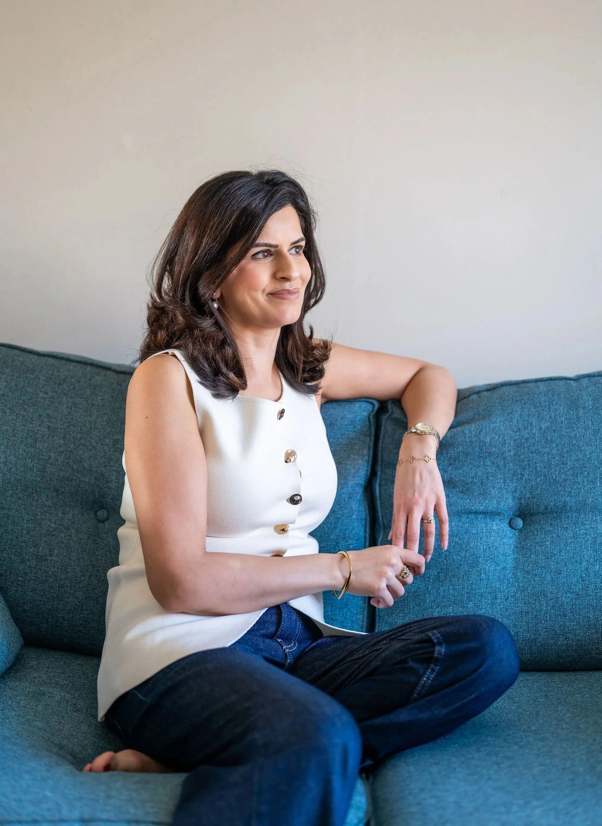 A woman with dark hair sits on a teal couch, wearing a sleeveless white top with buttons, dark jeans, and jewelry, looking slightly to the right.