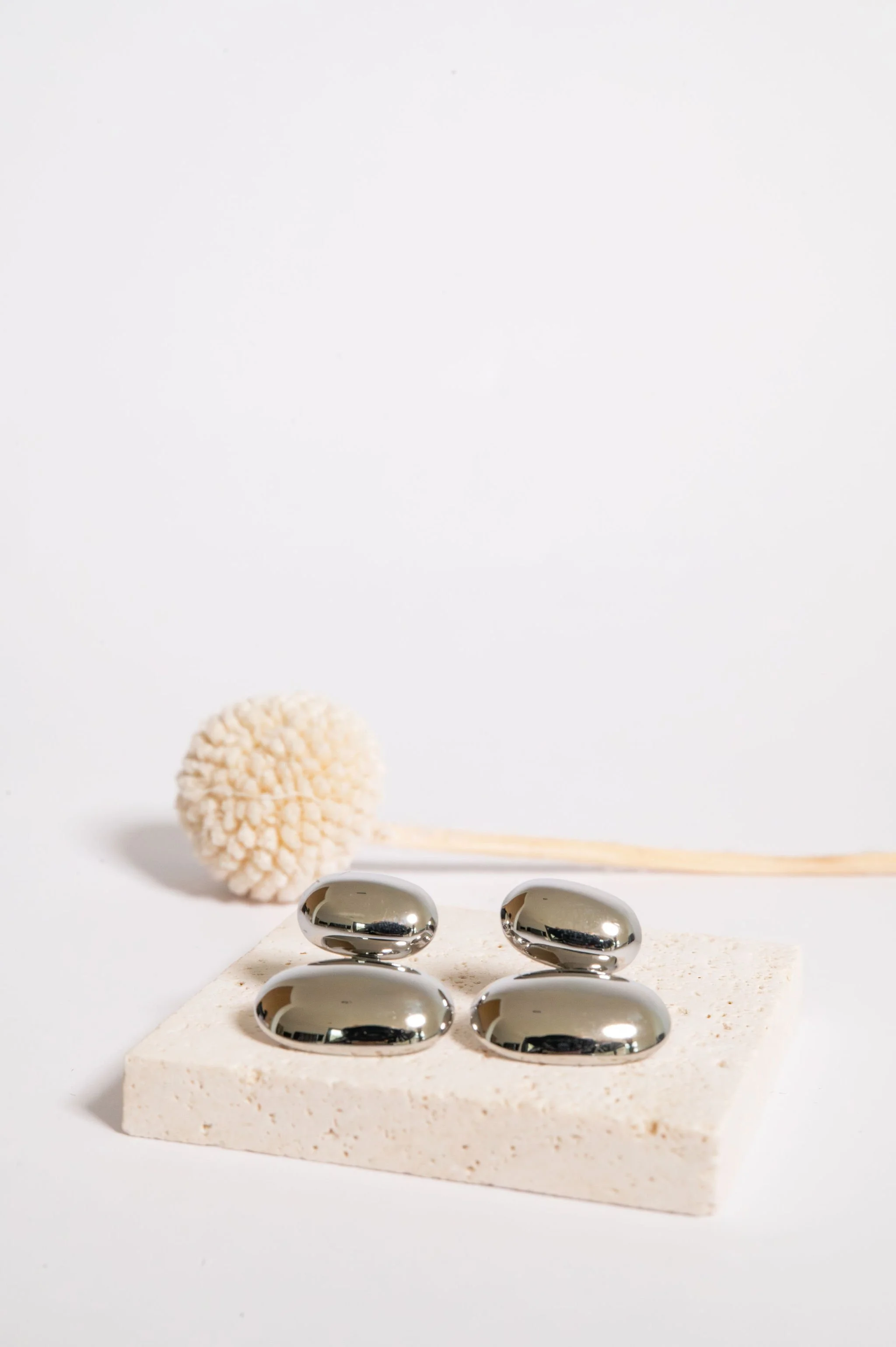 Four metallic, pebble-shaped objects arranged on a white textured platform with a beige pom-pom and a thin stick behind them, against a plain white background.