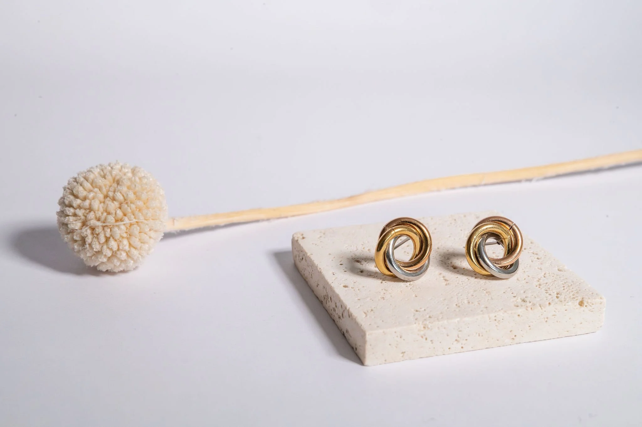 Pair of earrings, one gold and one silver, placed on a beige stone slab with a dried flower nearby on a white background.