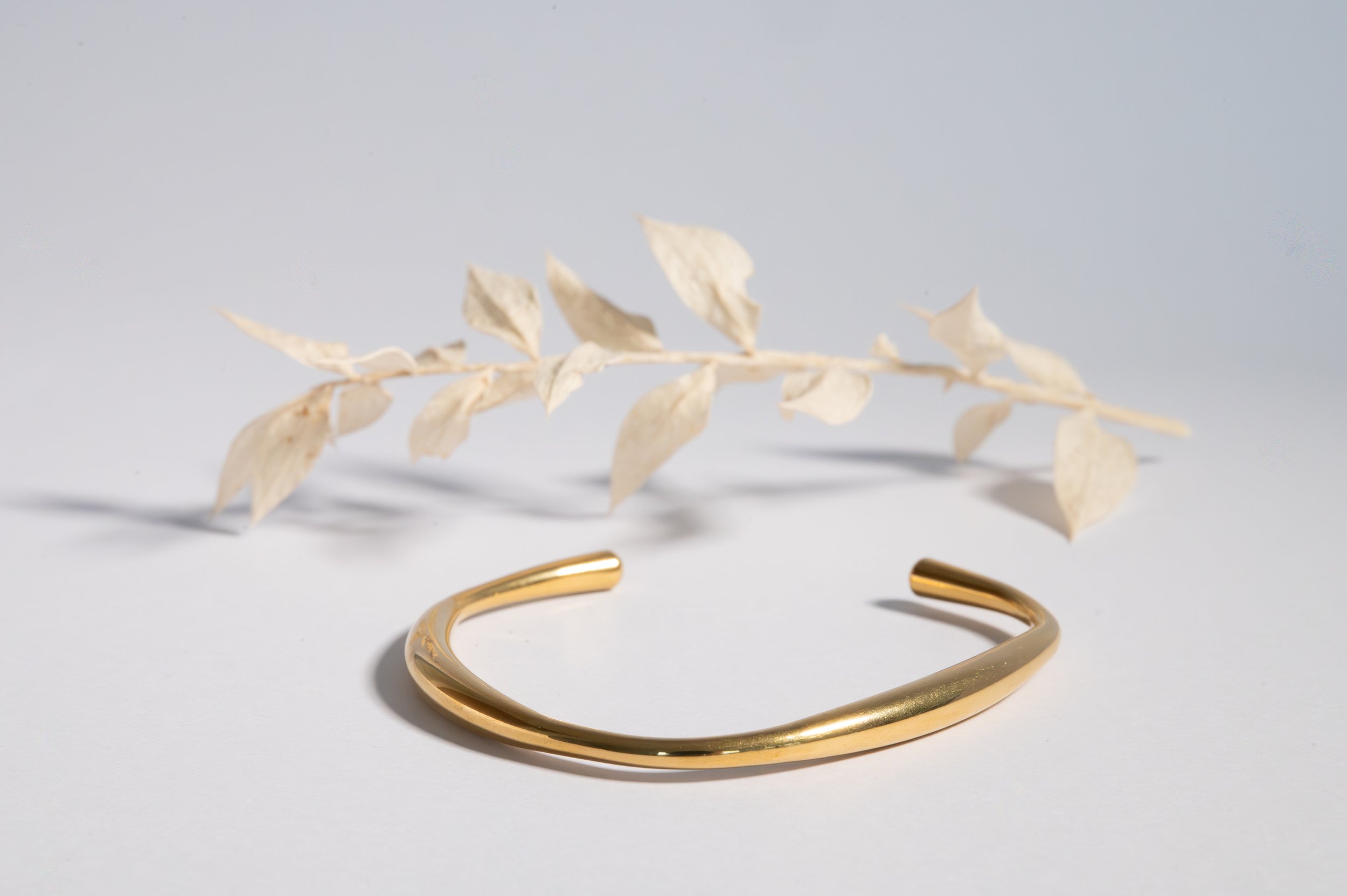 Gold cuff bracelet on white surface with dried beige leaves in the background.