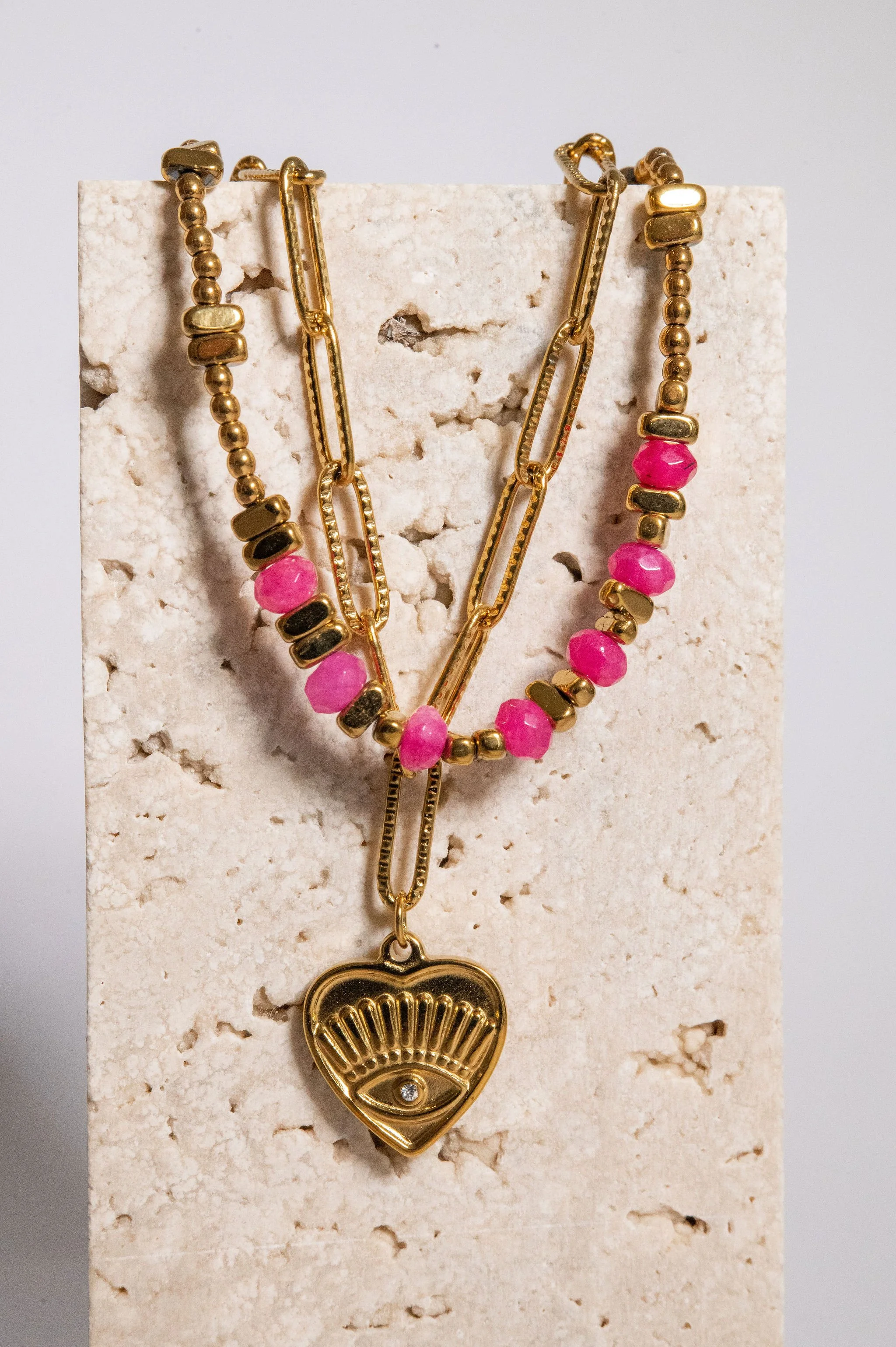 Close-up of layered gold necklaces with pink and black beads and a heart-shaped pendant displaying an eye design, hanging on a textured white surface.