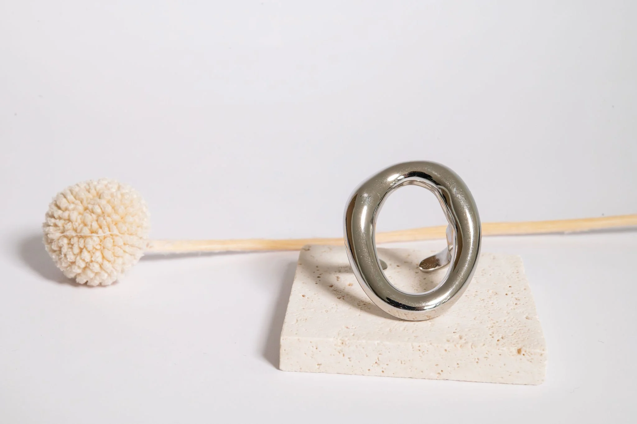 A metallic ring sculpture placed on a beige rectangular stone block with a white background, with a cotton swab with a fluffy tip laying horizontally behind it.