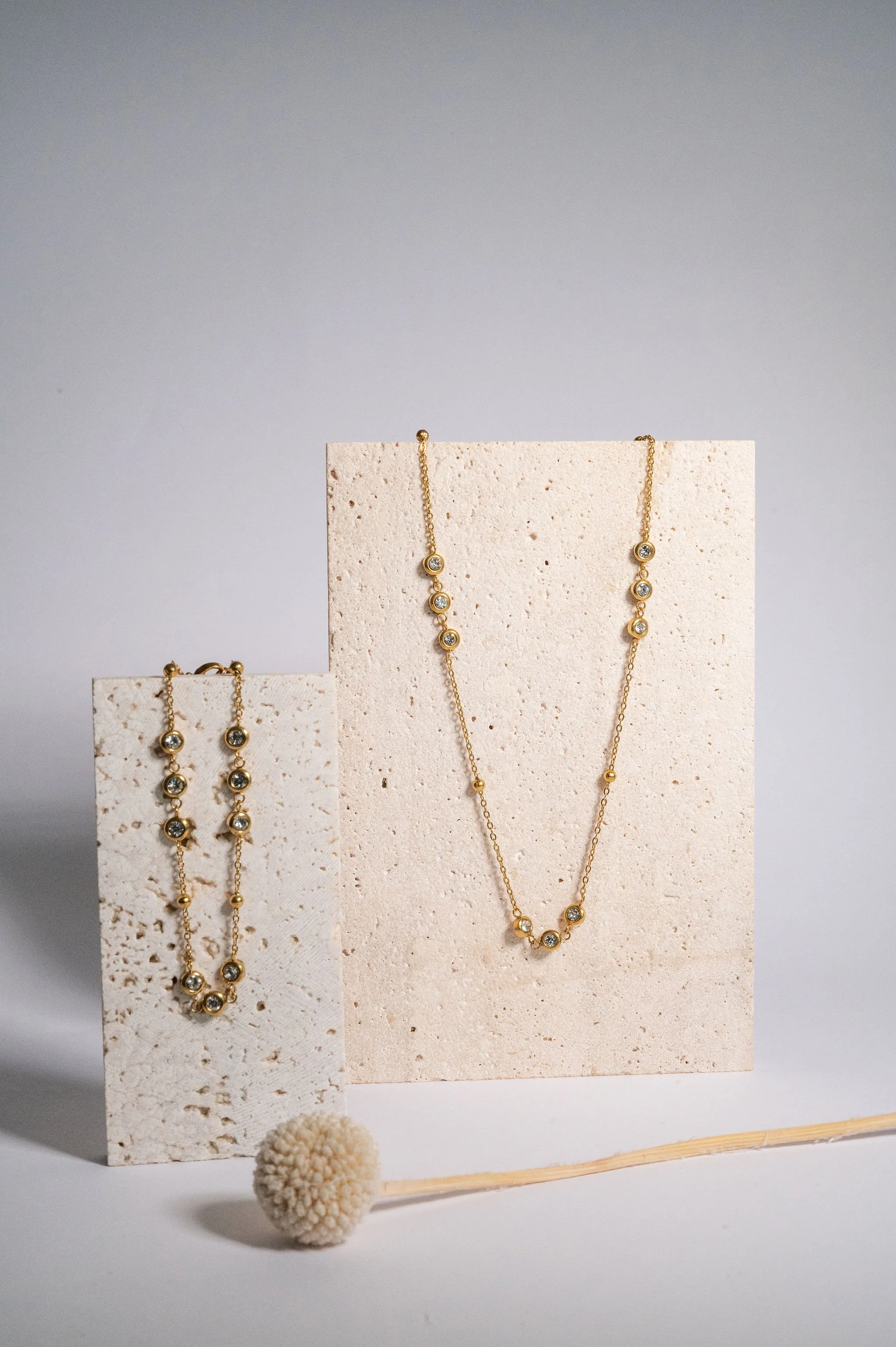 Gold jewelry set including a necklace and earrings with round gemstones arranged in a line on light-colored textured display blocks, with a small pom-pom on a stick in front.