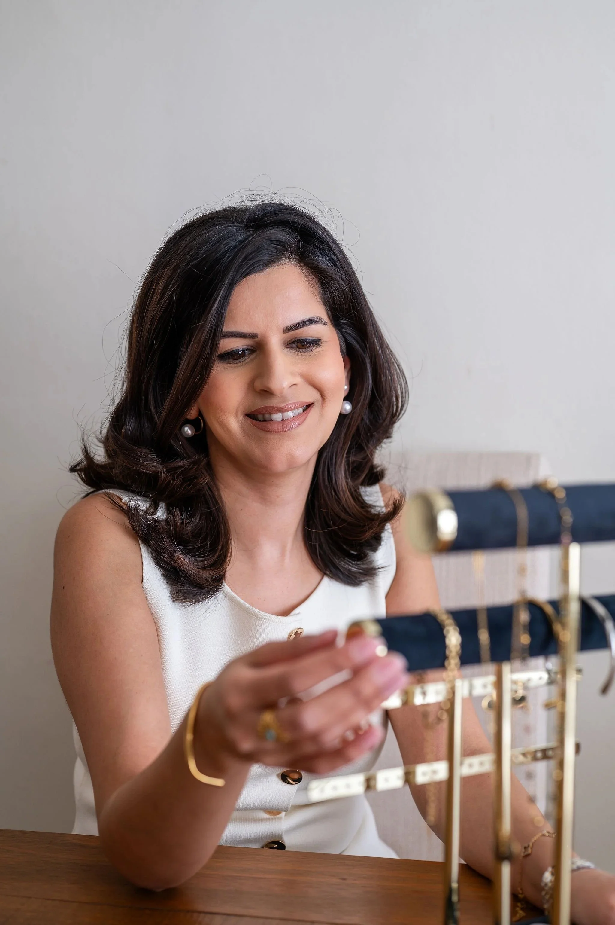 A woman with dark shoulder-length hair, wearing pearl earrings and a white sleeveless top, is smiling as she looks at jewelry displayed on a black and gold jewelry stand.