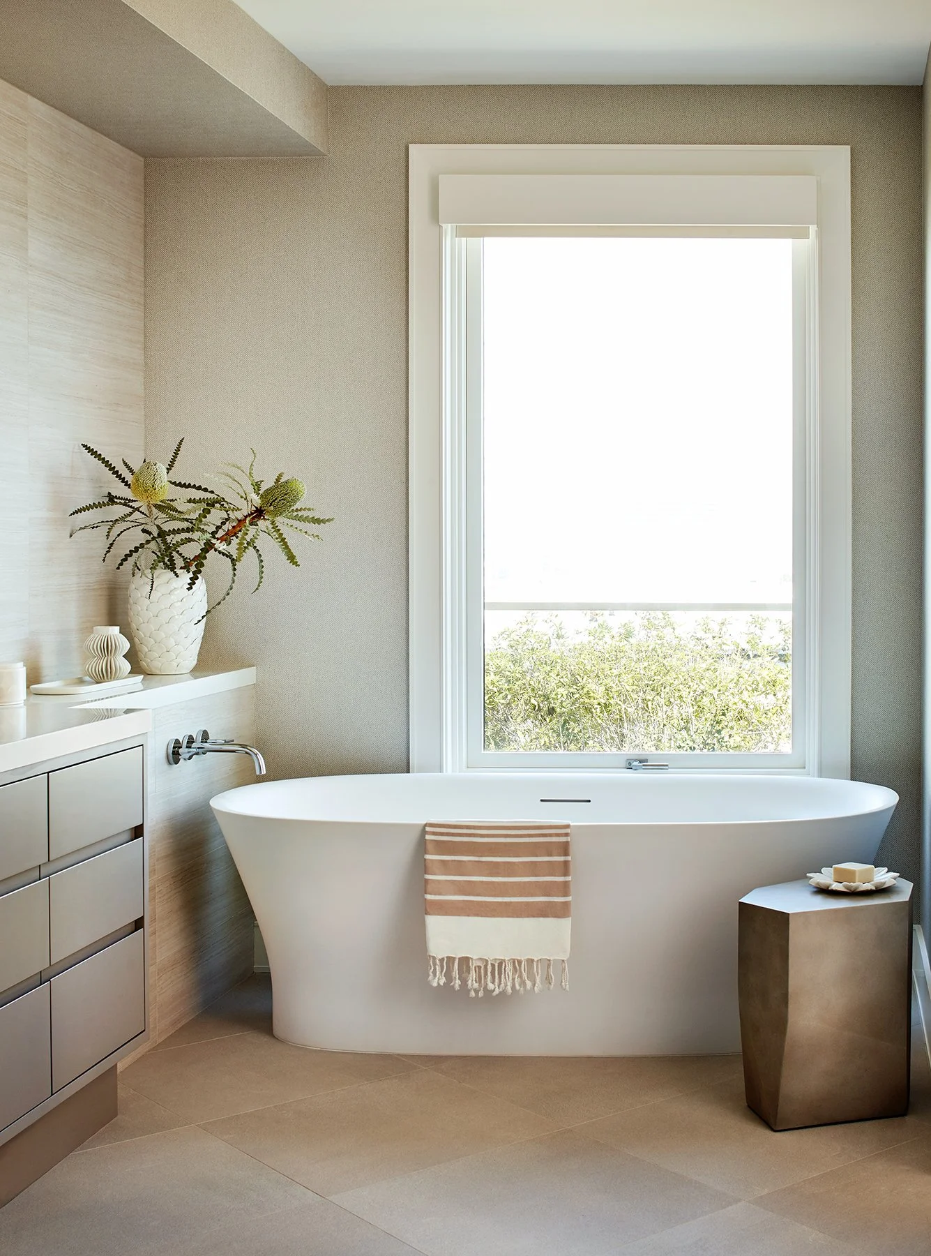 Modern bathroom with a freestanding bathtub, a window with a view of greenery, and minimal decor including a large plant, soap, and candles.