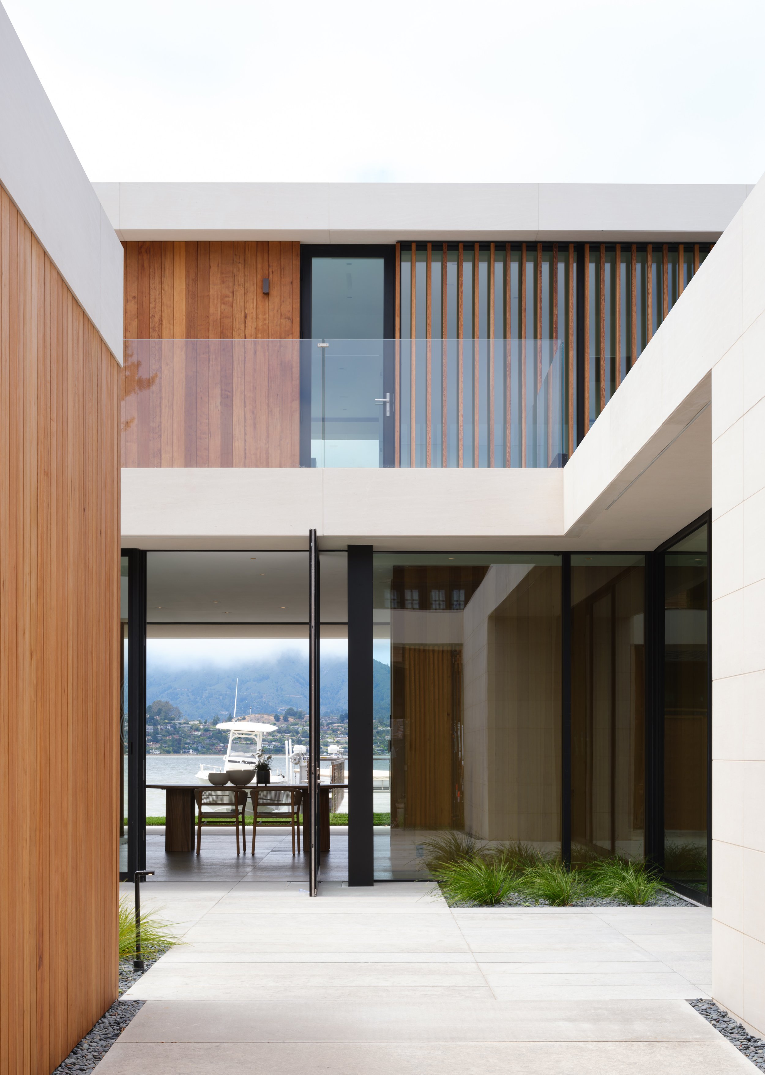 Modern Tiburon waterfront home in Marin County with wood, white, and glass exterior, overlooking the bay with a boat and distant hills, showcasing modern coastal architecture and indoor outdoor living.
