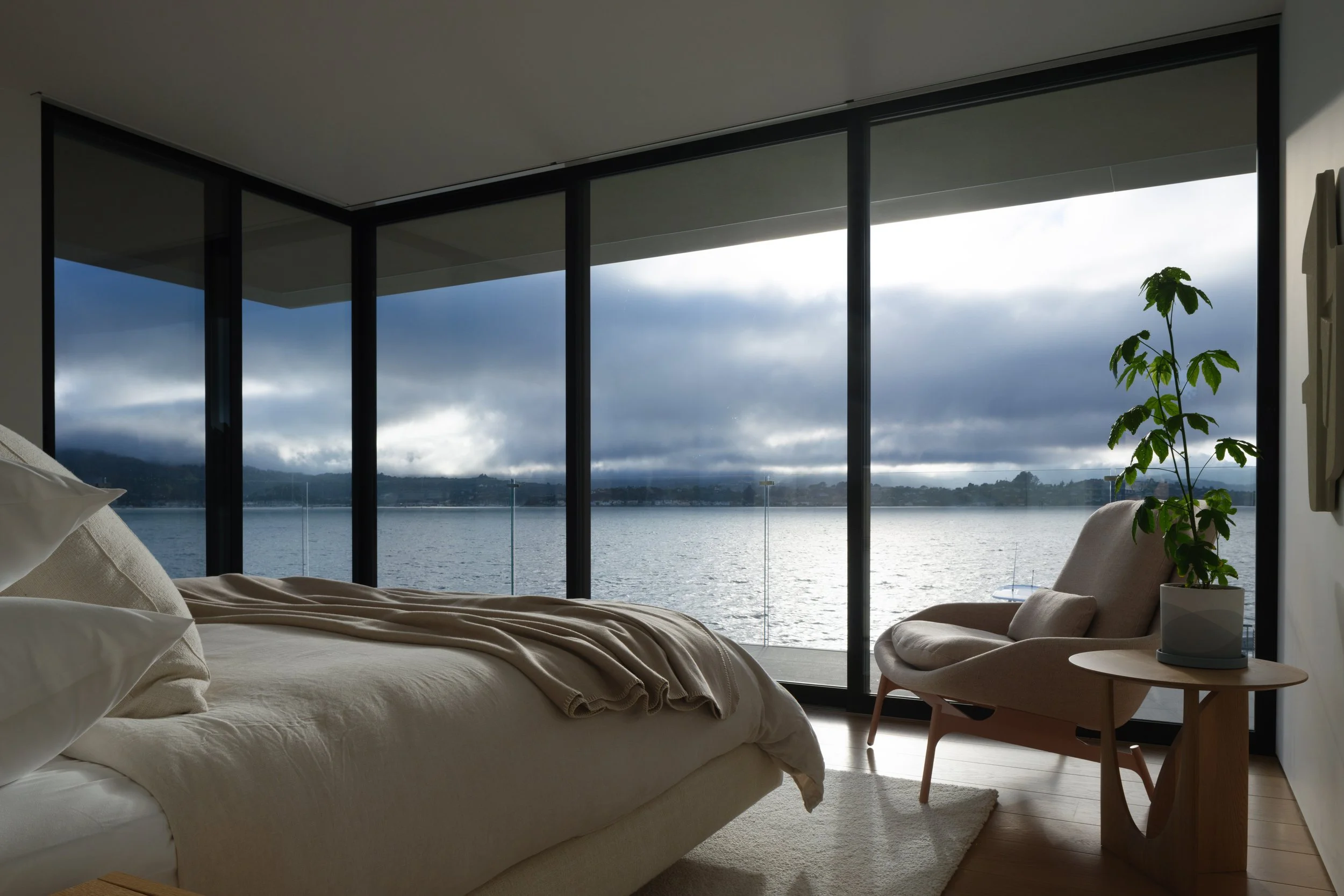 Tiburon waterfront bedroom with floor to ceiling windows overlooking the bay, styled with a simple bed, lounge chair, side table, and indoor plant for calm modern luxury in Marin County interior design.