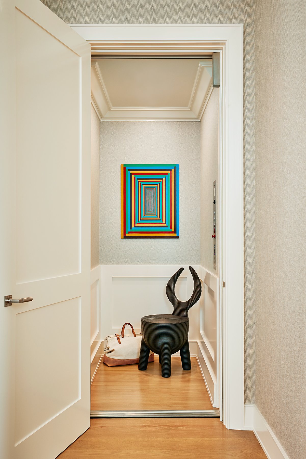 An elevator with white doors open, revealing a small interior decorated with a colorful geometric painting on the back wall, a black chair with curved horns, and a tote bag on the floor on Filbert Street Pacific Heights in San Francisco.