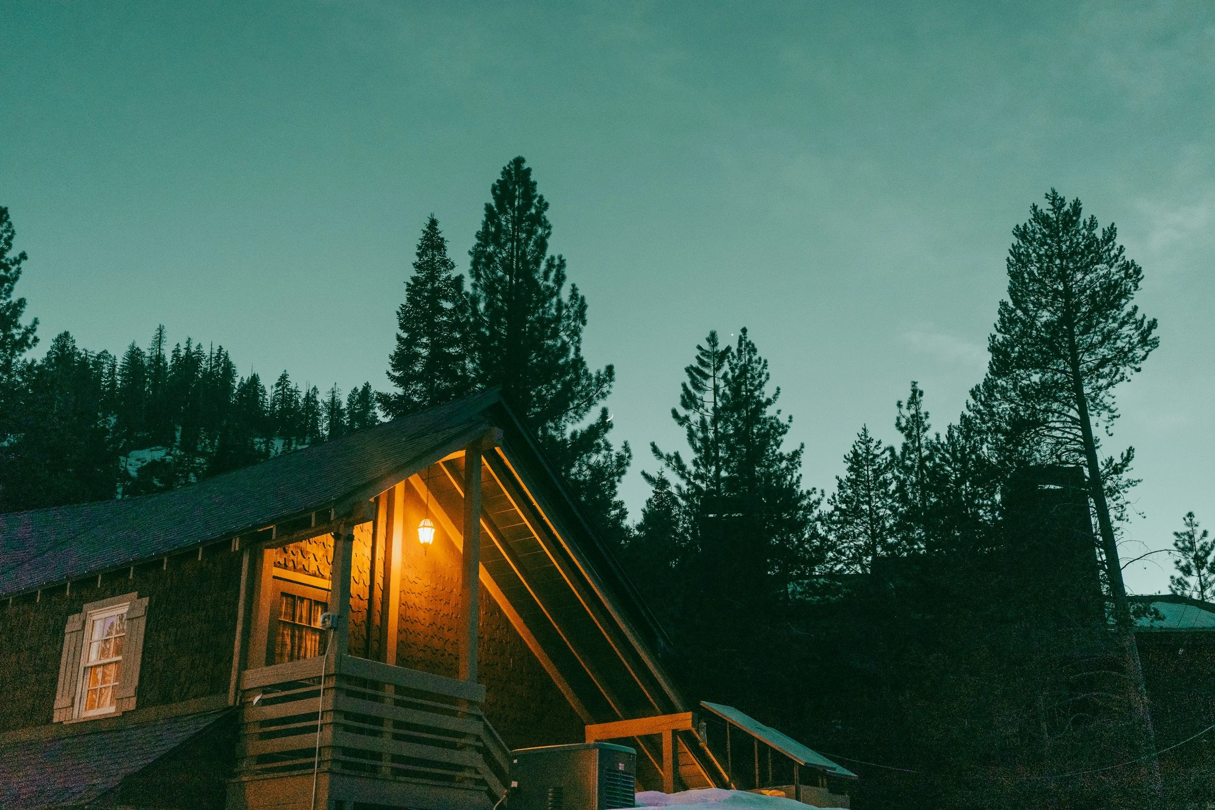 A house with a lit porch light surrounded by tall pine trees at dusk or early evening in Lake Tahoe