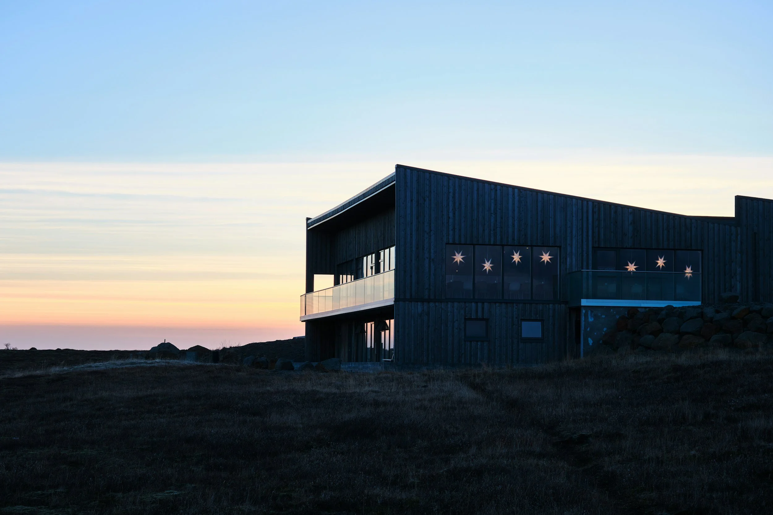 Modern house with wooden exterior and star-shaped lights in the windows, situated on a hill during sunset.  BallonStudio is a luxury interior design firm committed to sustainability in interior architecture in Sea Ranch