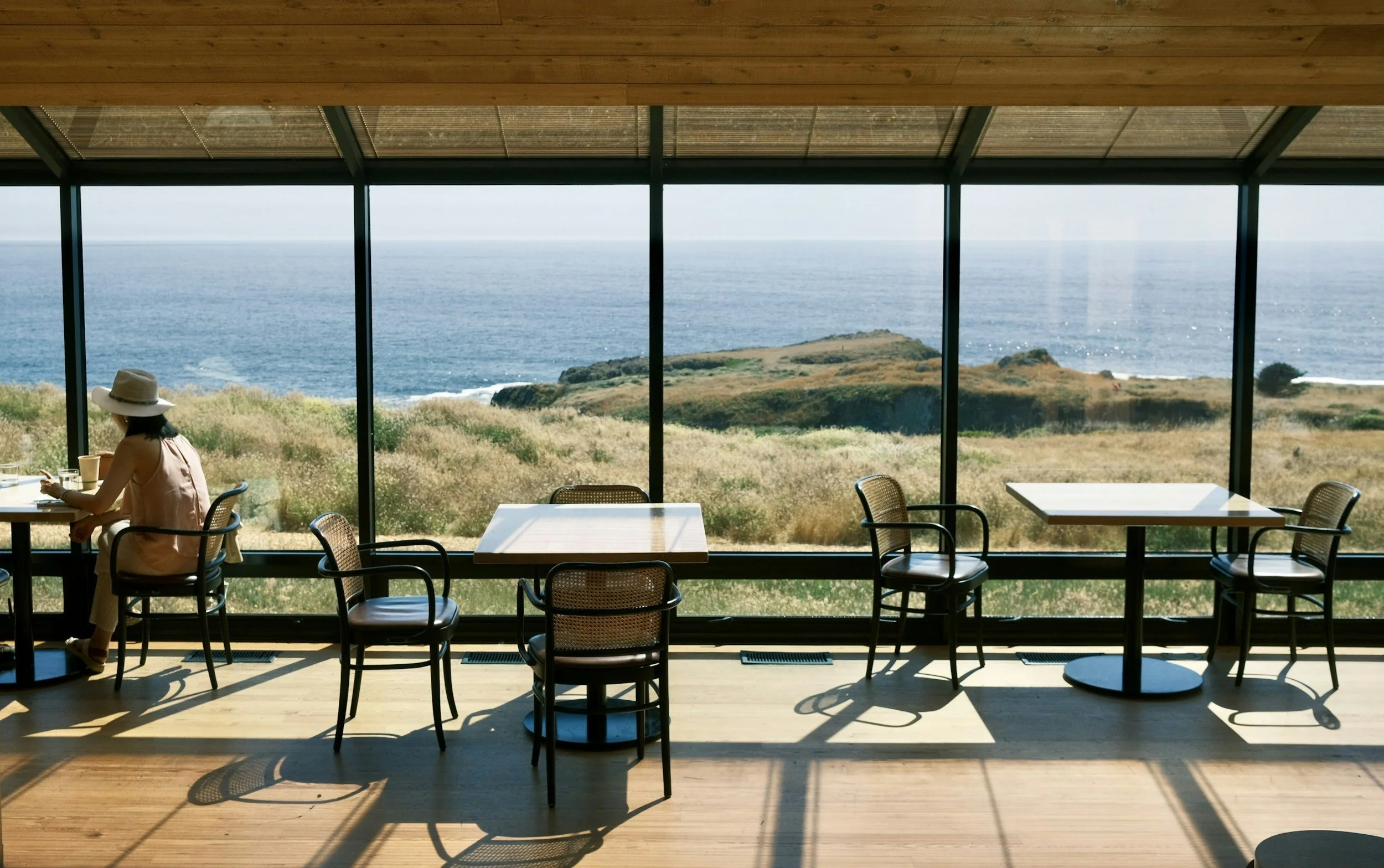 A woman sitting at a table in a cafe with a large glass window overlooking the coastline, grassy hills, and the ocean during daytime. BallonStudio is a luxury interior design firm committed to sustainability in interior architecture in Sea Ranch