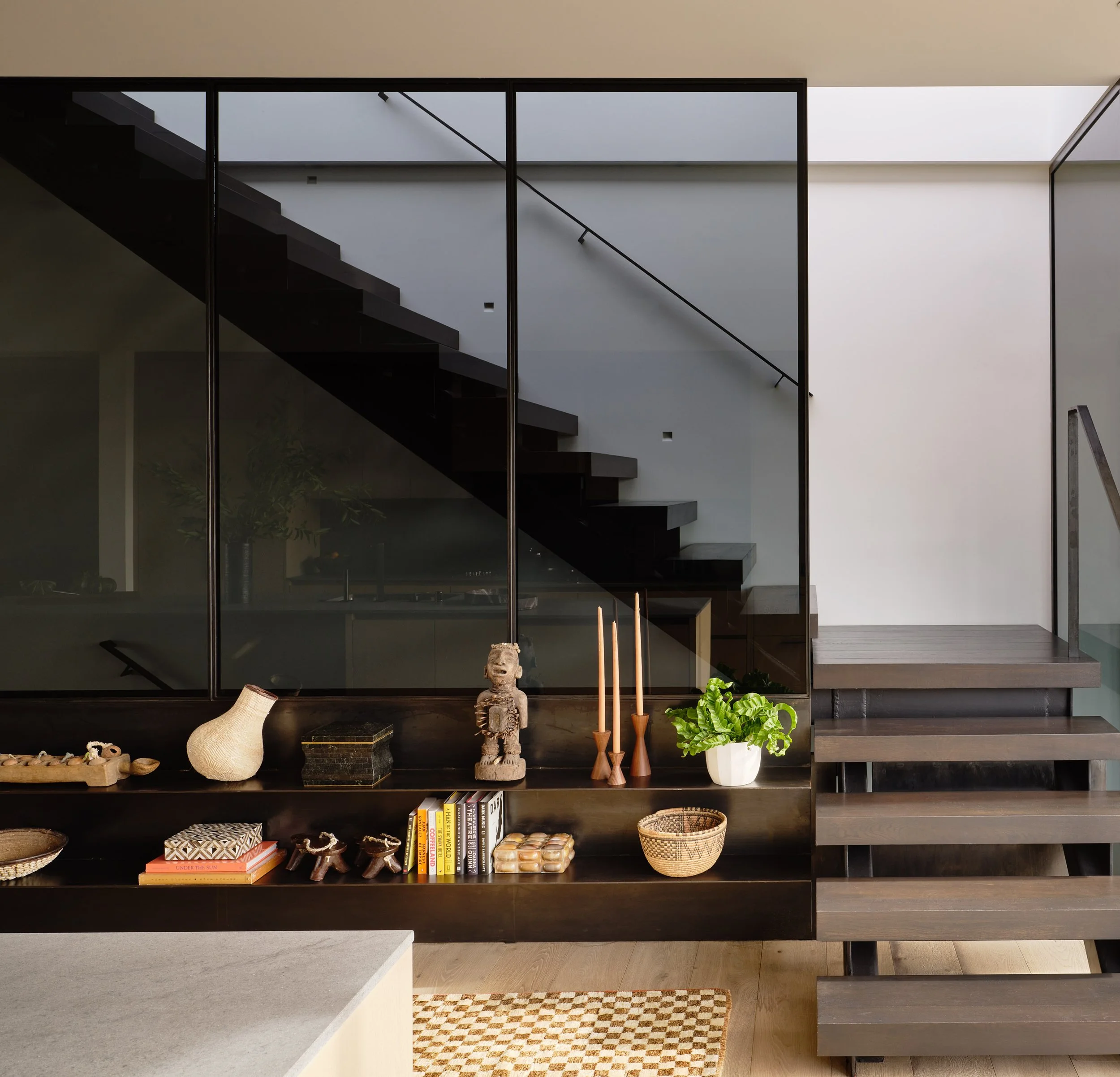 Broadway Penthouse Pacific Heights staircase with dark wood treads and glass railing, overlooking built in shelving styled with books, plants, candles, and art objects for curated modern luxury in San Francisco.