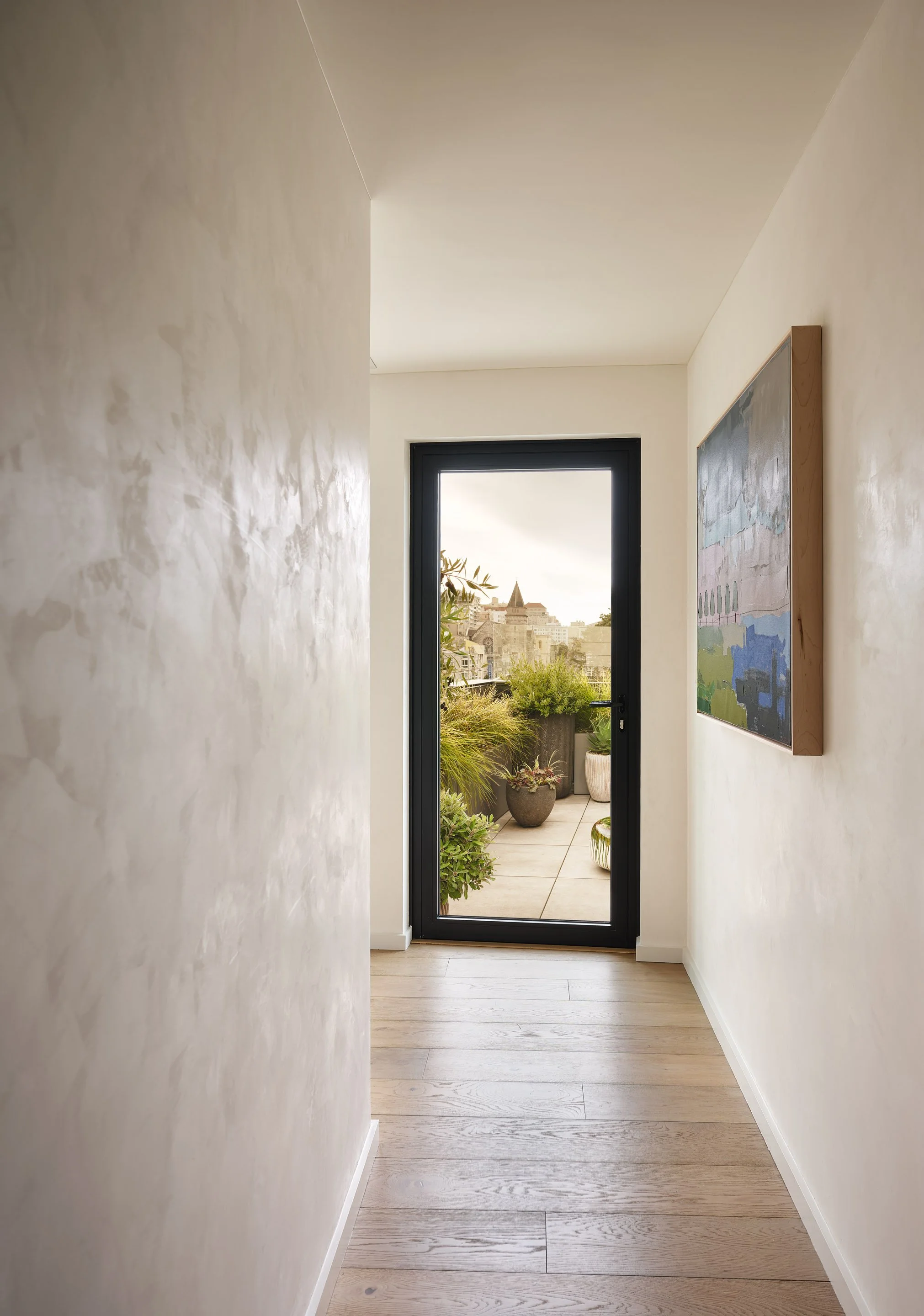 Broadway Penthouse Pacific Heights hallway with warm wood floors and framed art leading to a glass door and balcony with planters, opening to San Francisco skyline views for calm modern luxury living.