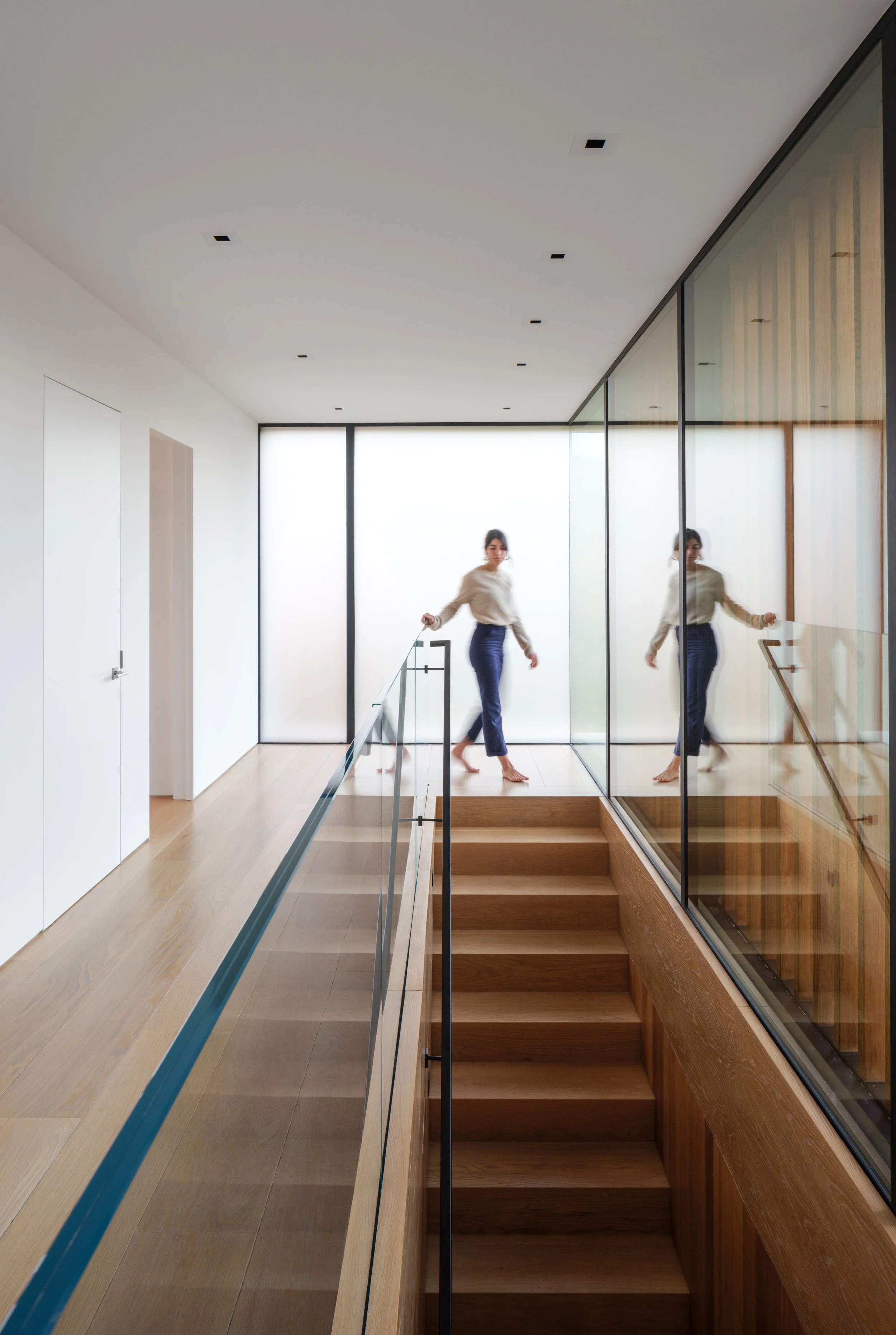 Minimalist Tiburon waterfront hallway with pale wood floors, glass stair railing, and floor to ceiling windows, showing modern Marin County interior design with calm indoor outdoor flow as a woman walks barefoot.