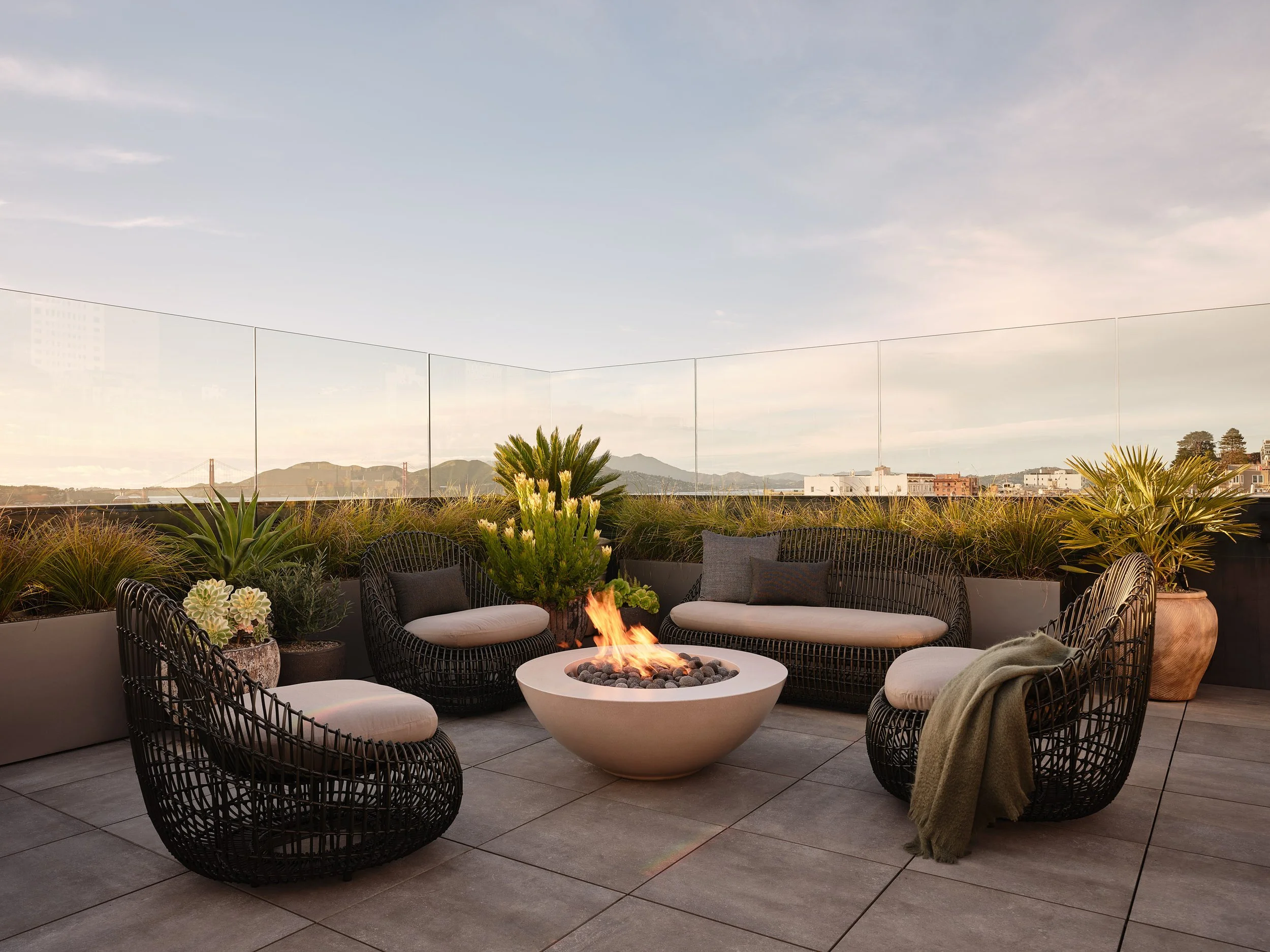 Broadway Penthouse Pacific Heights rooftop patio with black woven seating around a central fire pit, styled with potted plants and glass railing, overlooking San Francisco skyline views for modern luxury outdoor living.