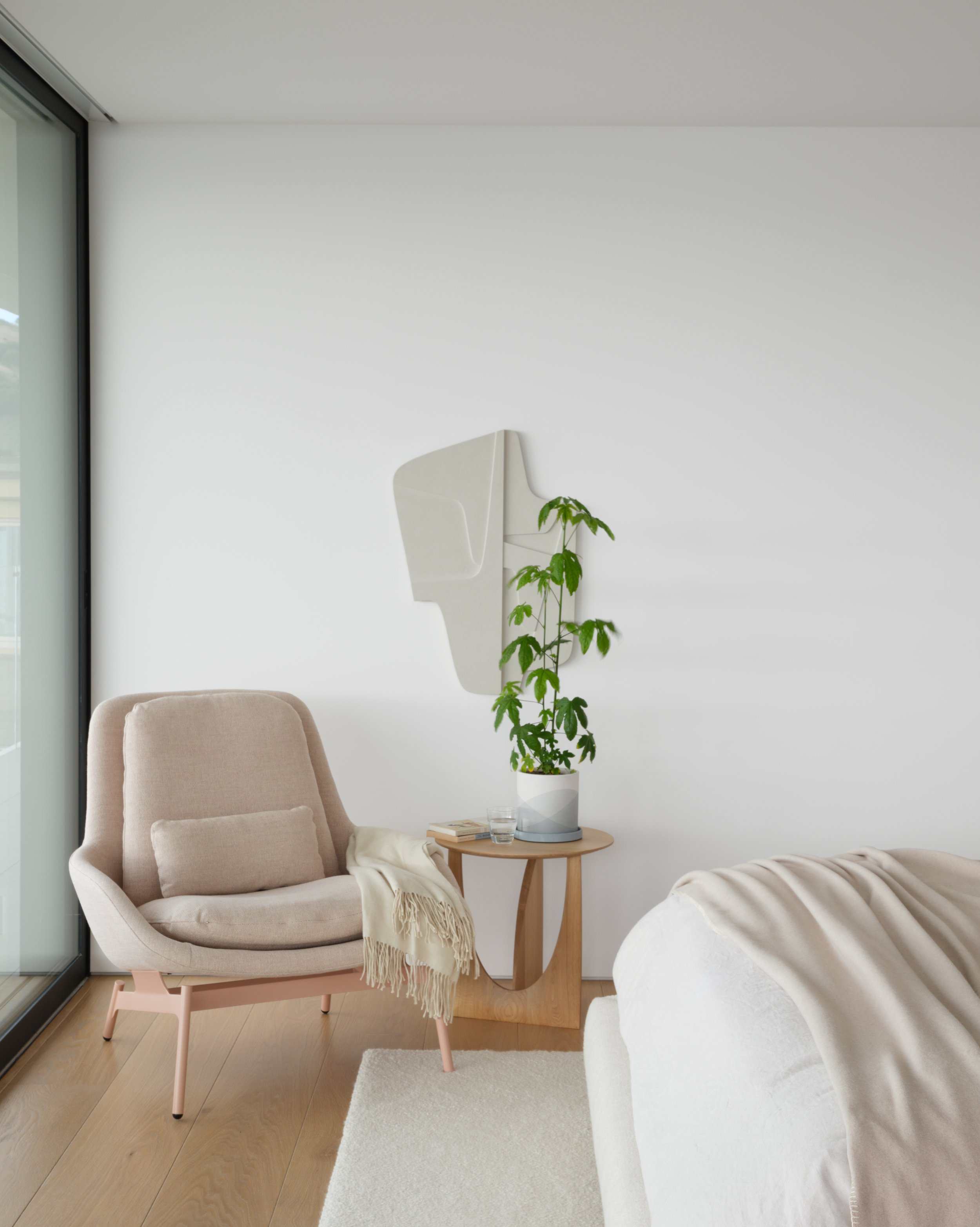 Tiburon waterfront living room with beige armchair, round wood side table with books and water, tall potted plant, and abstract art beside sliding glass doors for modern Marin County indoor outdoor living.