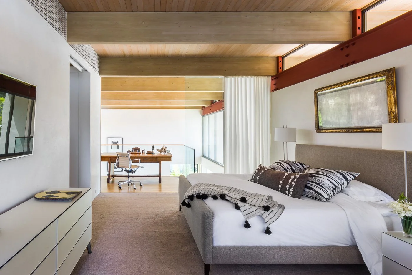 St. Helena Napa Valley bedroom and workspace with layered bedding beside a desk and office chair, framed by large windows and exposed wood ceiling beams for calm luxury living in a wine country retreat.