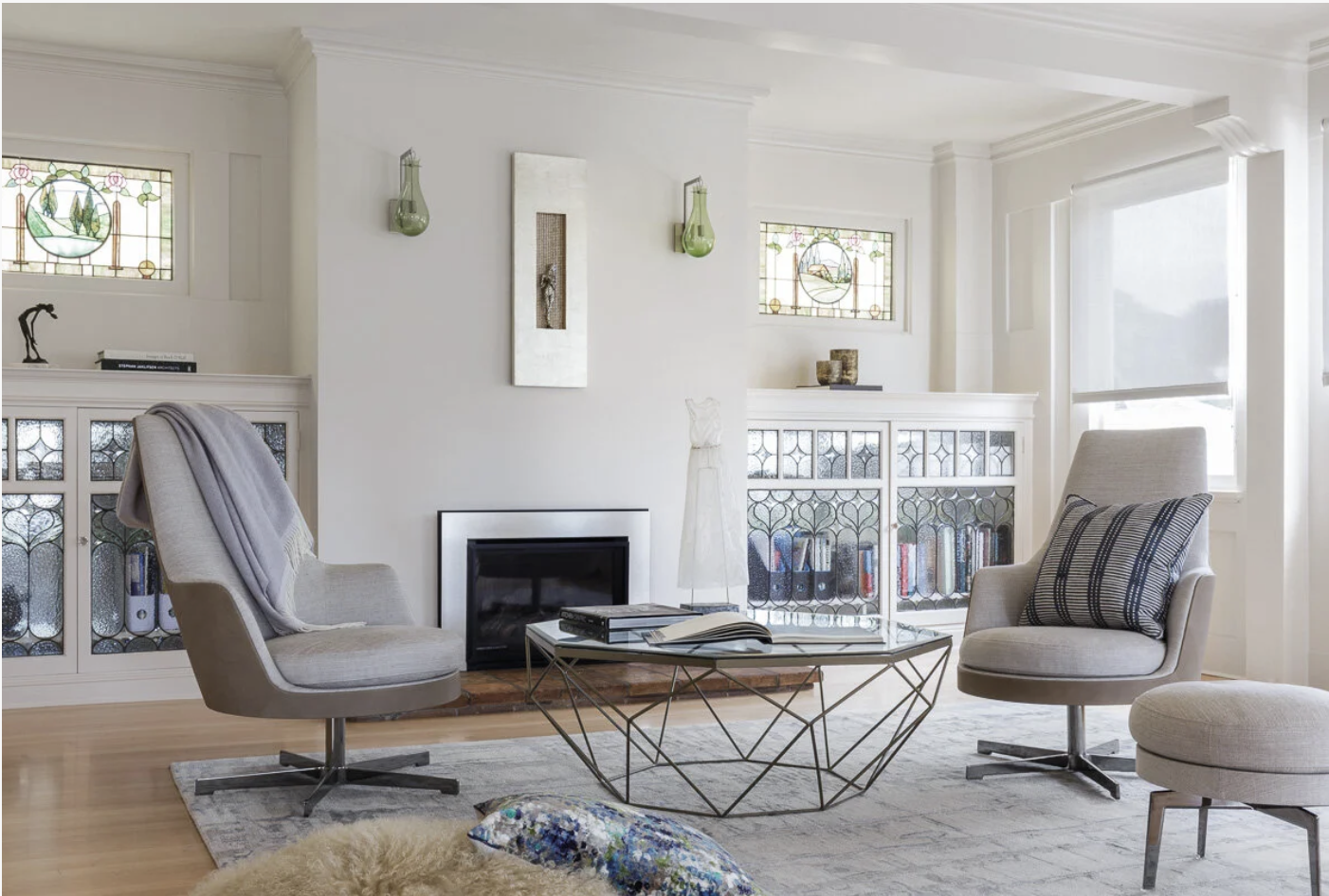 Home office lounge with two beige armchairs and a glass coffee table, centered on a fireplace and built in bookshelves, framed by stained glass windows and soft curtains for calm luxury residential living.