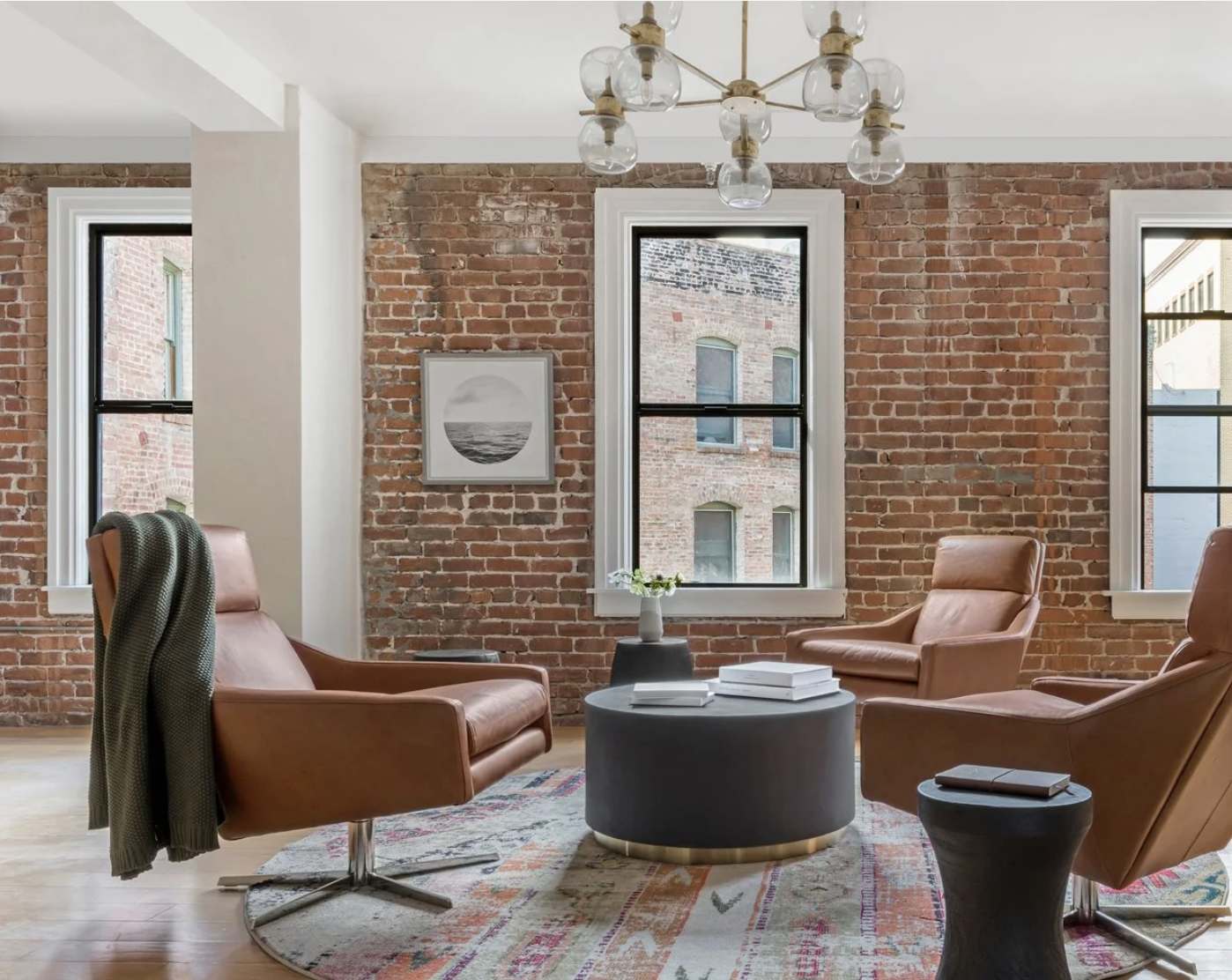 Office lounge with exposed brick walls and three large windows, featuring modern leather chairs around a coffee table, a glass globe chandelier overhead, and framed artwork creating a warm, refined gathering space.