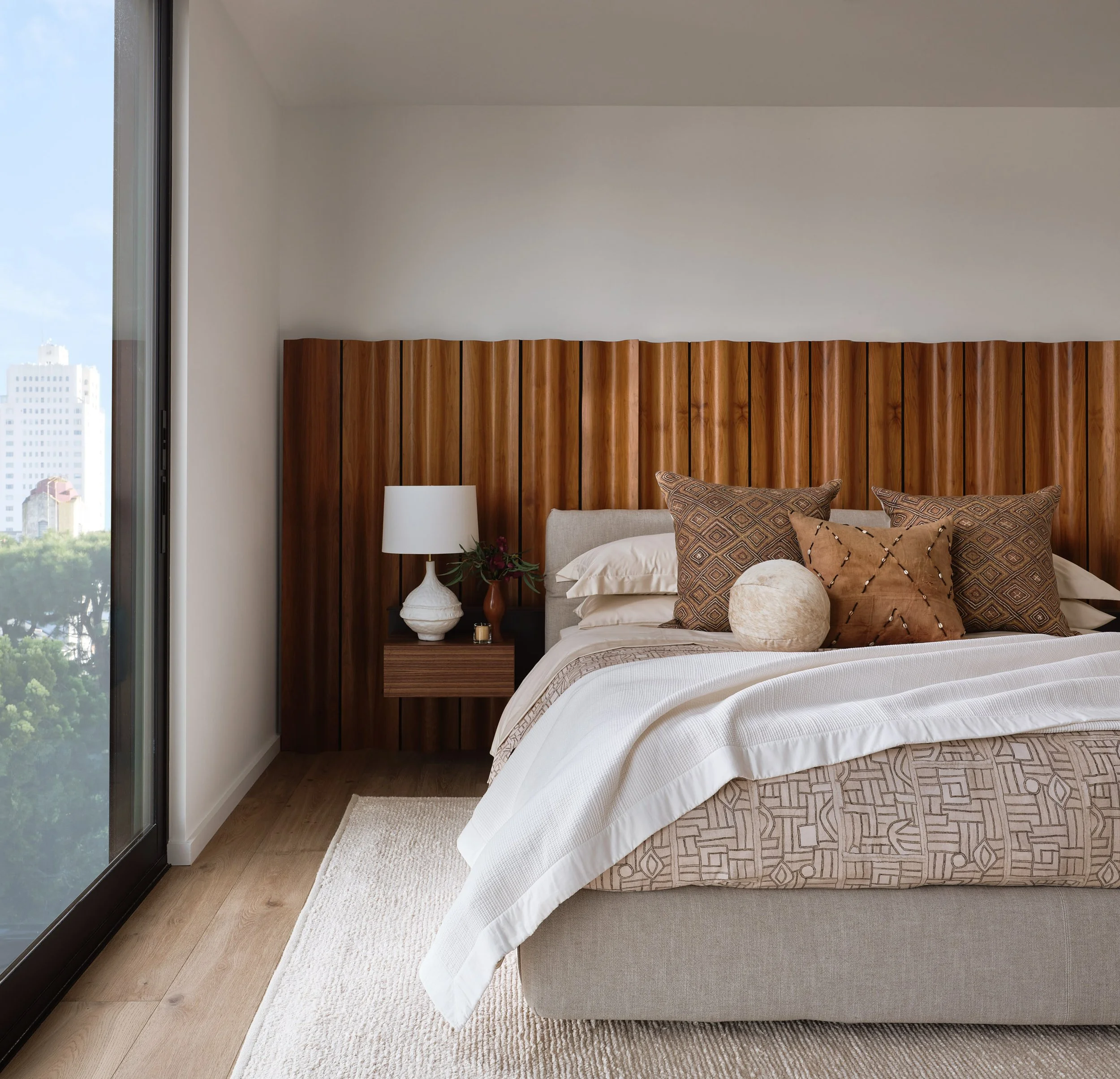 Broadway Penthouse Pacific Heights primary bedroom with a warm wood headboard and layered neutral bedding, styled with a sculptural lamp and greenery beside a large window with San Francisco city views.