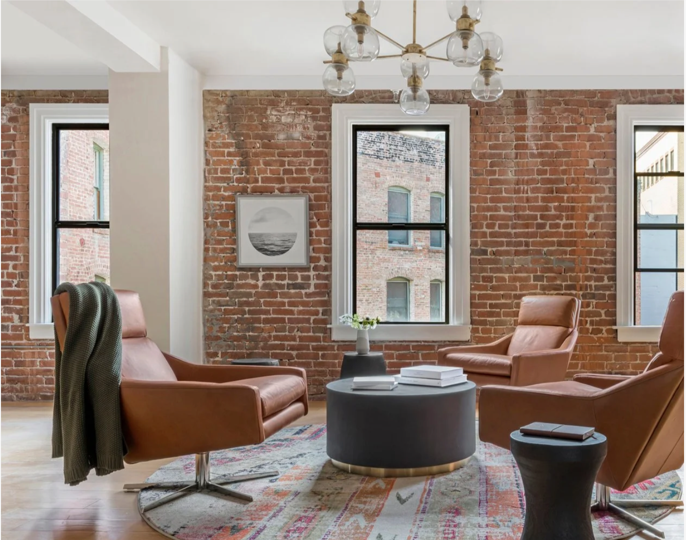 Tech company workspace with conformable living room meeting lounge with an exposed brick wall and three large windows, styled with mid century chairs, a round coffee table, and a glass globe chandelier for warm modern luxury living.