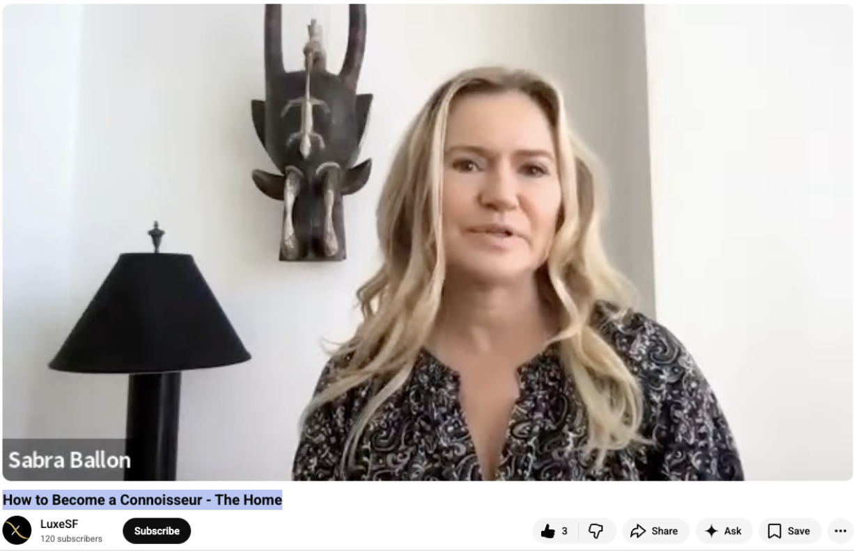 A woman with long blonde hair wearing a black and white patterned top speaking in a video titled "How to Become a Connoisseur - The Home". Behind her is a black lamp and a wooden African tribal mask on the wall.
