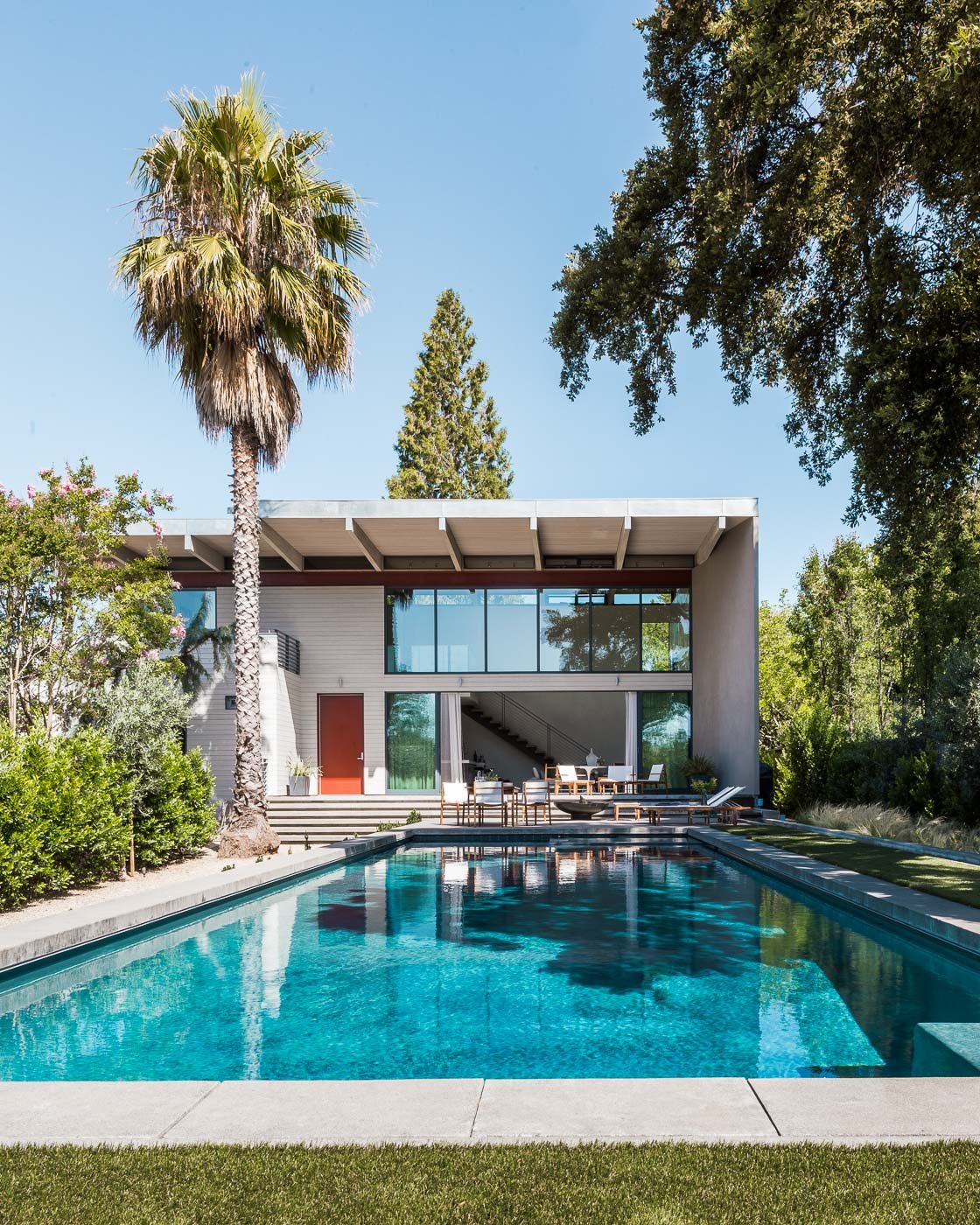 St. Helena Napa Valley modern home with large glass windows opening to outdoor seating and a backyard swimming pool, surrounded by lush greenery under a clear sky for relaxed luxury indoor outdoor living.