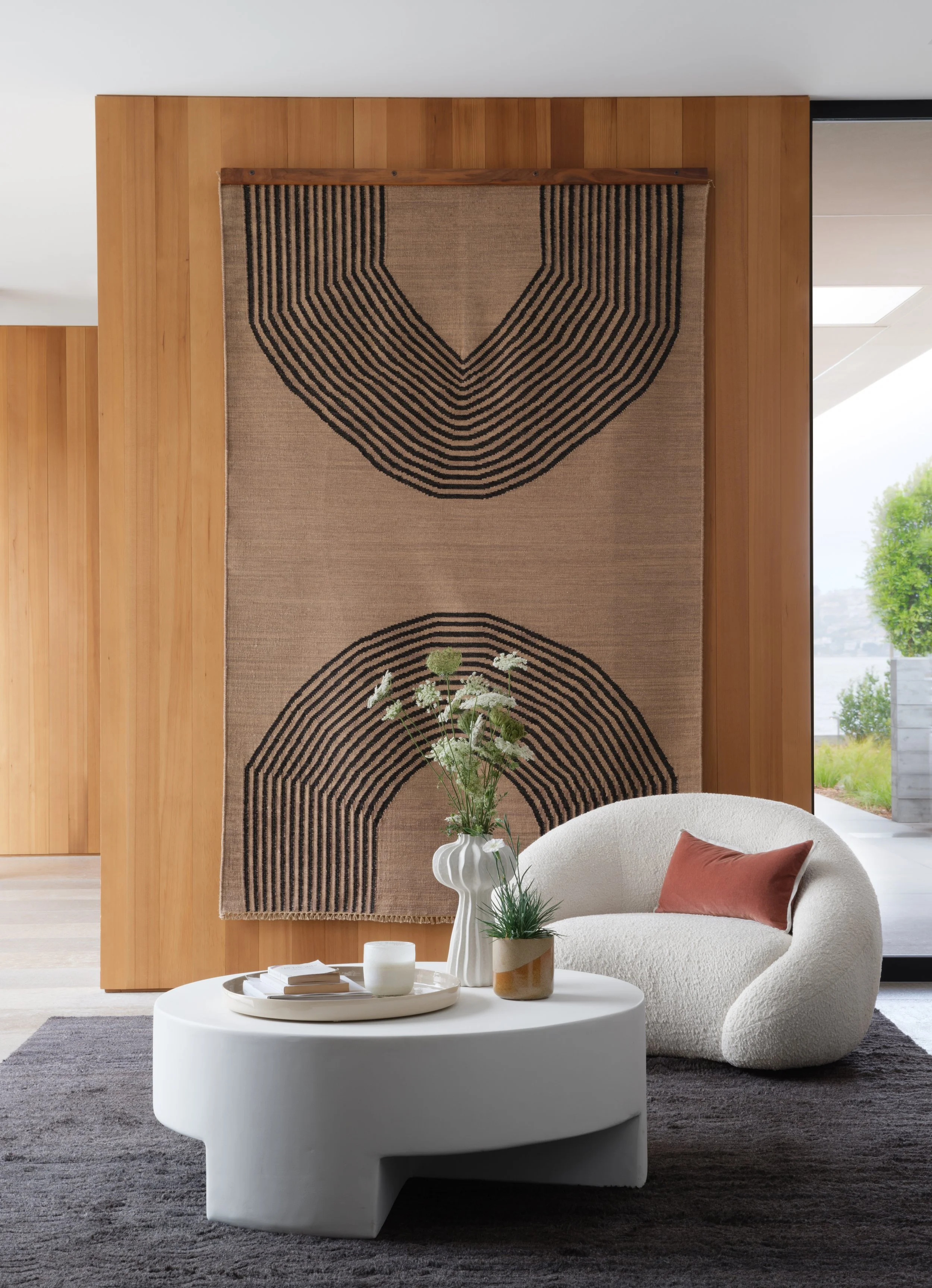 Tiburon waterfront living room with beige textured lounge chair and round white coffee table, styled with flowers and a large geometric textile artwork, framed by warm wood walls and glass for modern Marin County interior design.