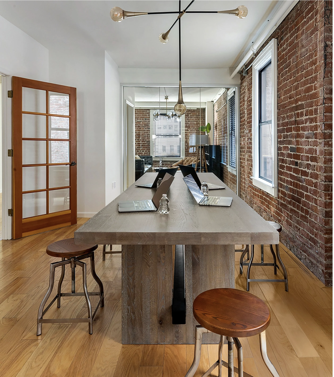 Geometer tech company conference room with exposed brick walls and a large wood table set with laptops and water bottles, surrounded by mixed metal stools and natural light from tall windows.