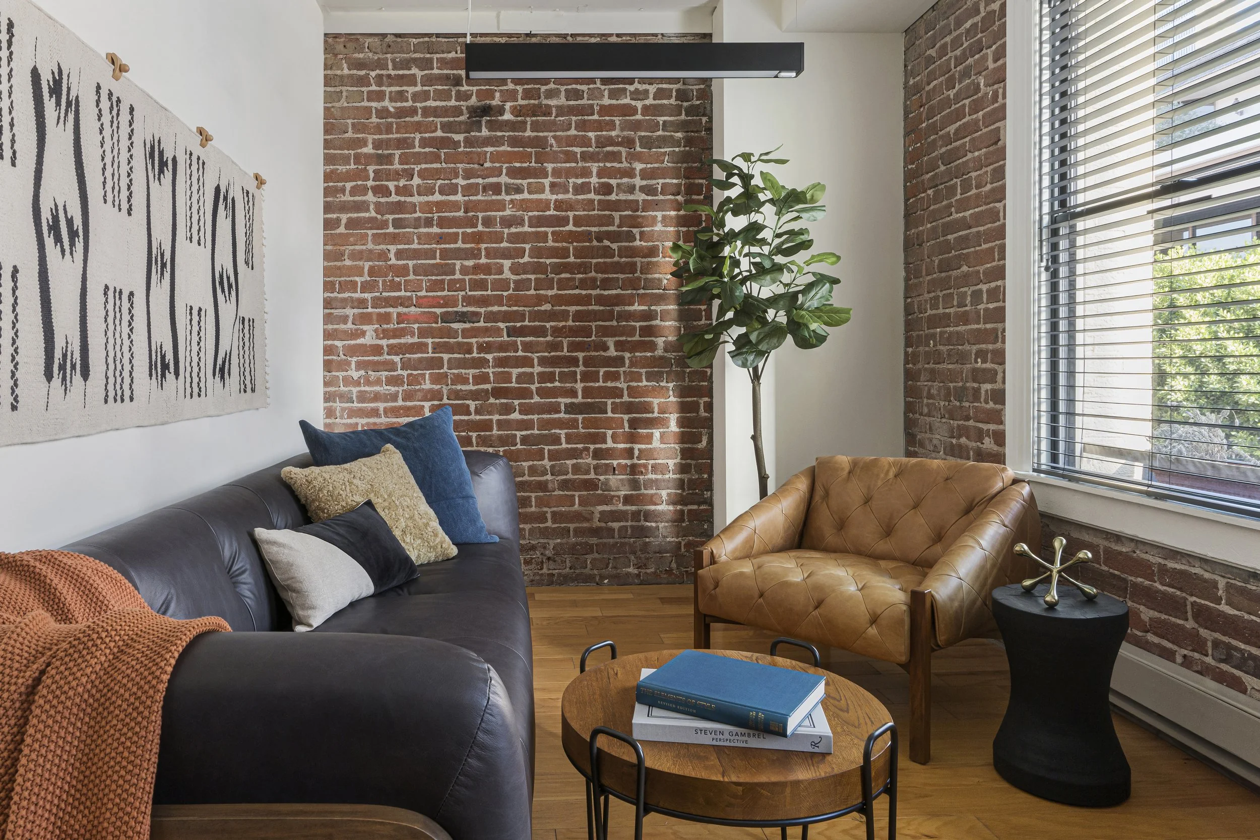 Office lounge with exposed brick wall and large window blinds, featuring a gray leather sofa with cushions, wood coffee table with books, tan leather armchair, black side table, and tall plant for warm modern workspace comfort.