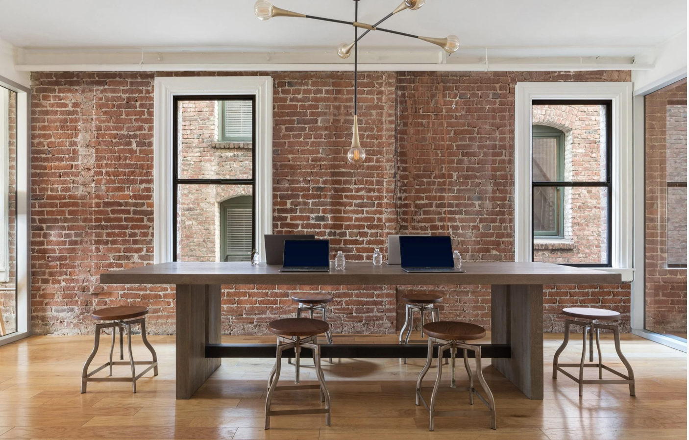 Modern conference room with a large wood table set with laptops and six stools, framed by exposed brick walls and tall windows, creating a bright, collaborative meeting space in a tech office.