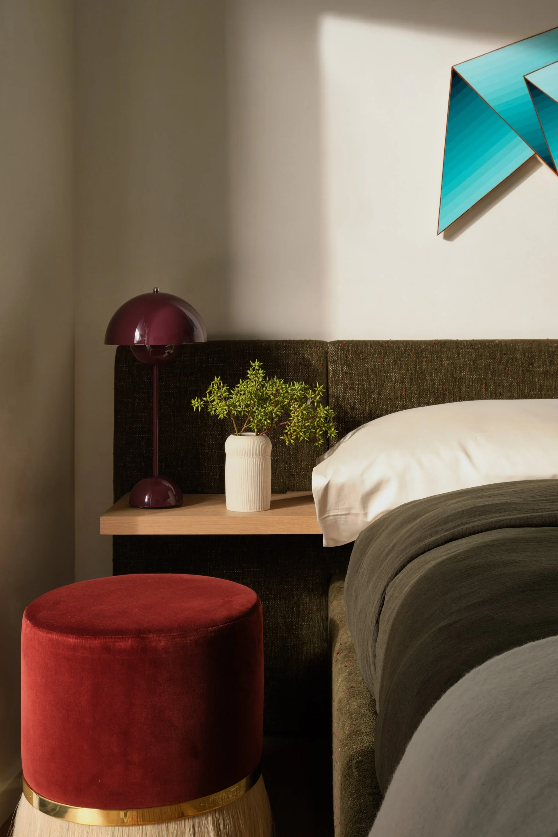 Broadway Penthouse Pacific Heights bedroom vignette with a dark upholstered headboard, maroon lamp, red velvet stool, and bedside styling with greenery and abstract art, lit by warm sun for calm modern luxury in San Francisco.