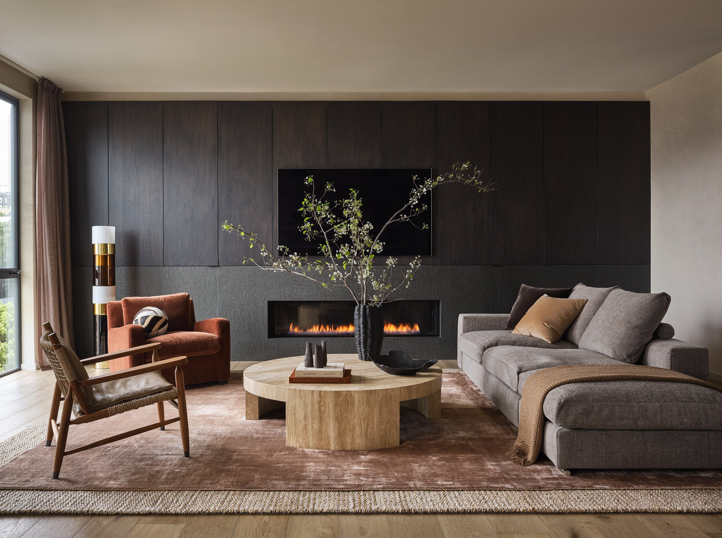 Pacific Heights Broadway penthouse living room with gray sectional and rust lounge chair, styled around a round wood coffee table, textured rug, and sleek fireplace set in dark wood paneling beside large windows with soft drapery.