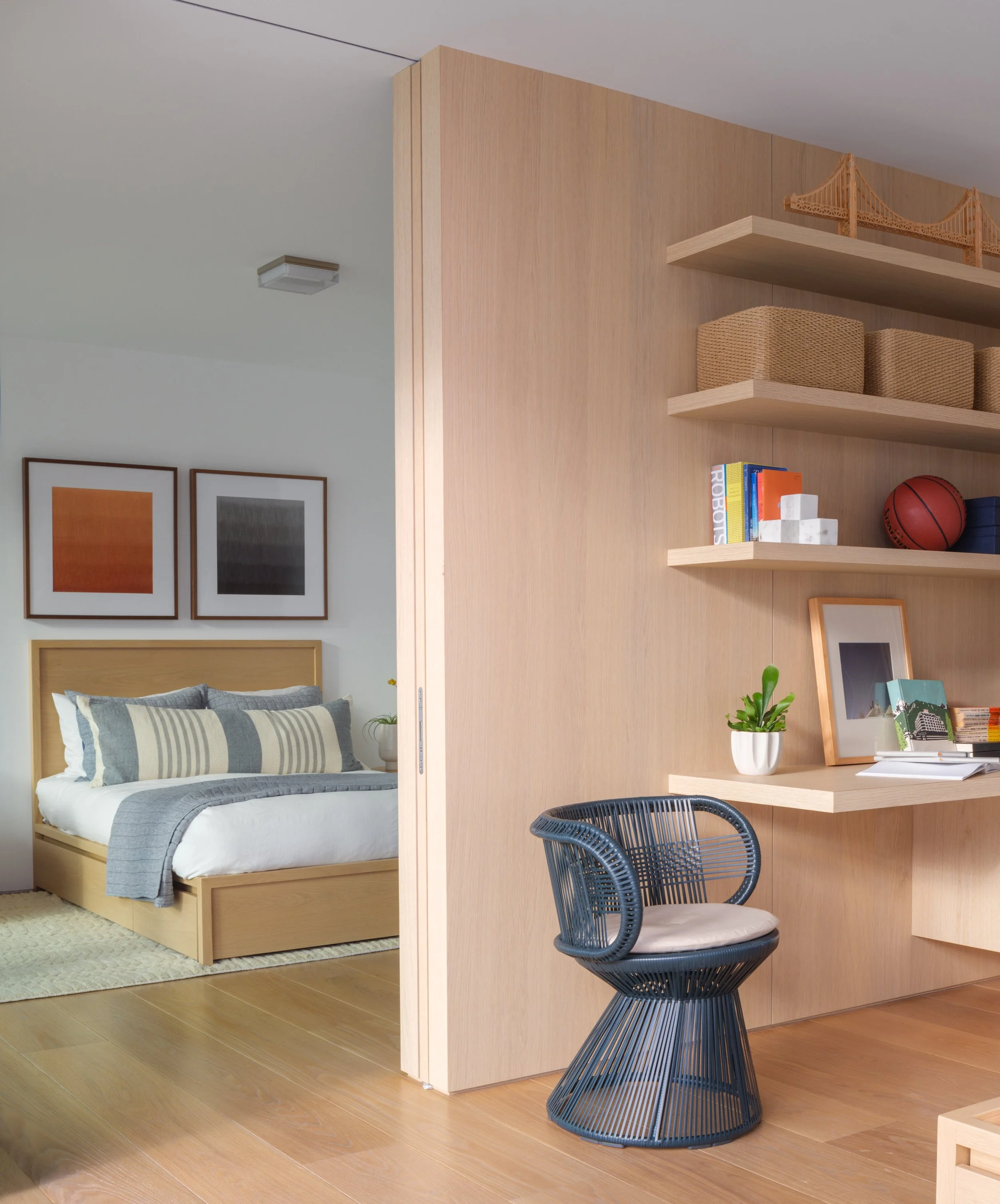 Tiburon waterfront bedroom and workspace with warm wood furniture, styled bed, lounge chair, and built in shelving with books and decor, creating calm modern luxury for a Marin County home.