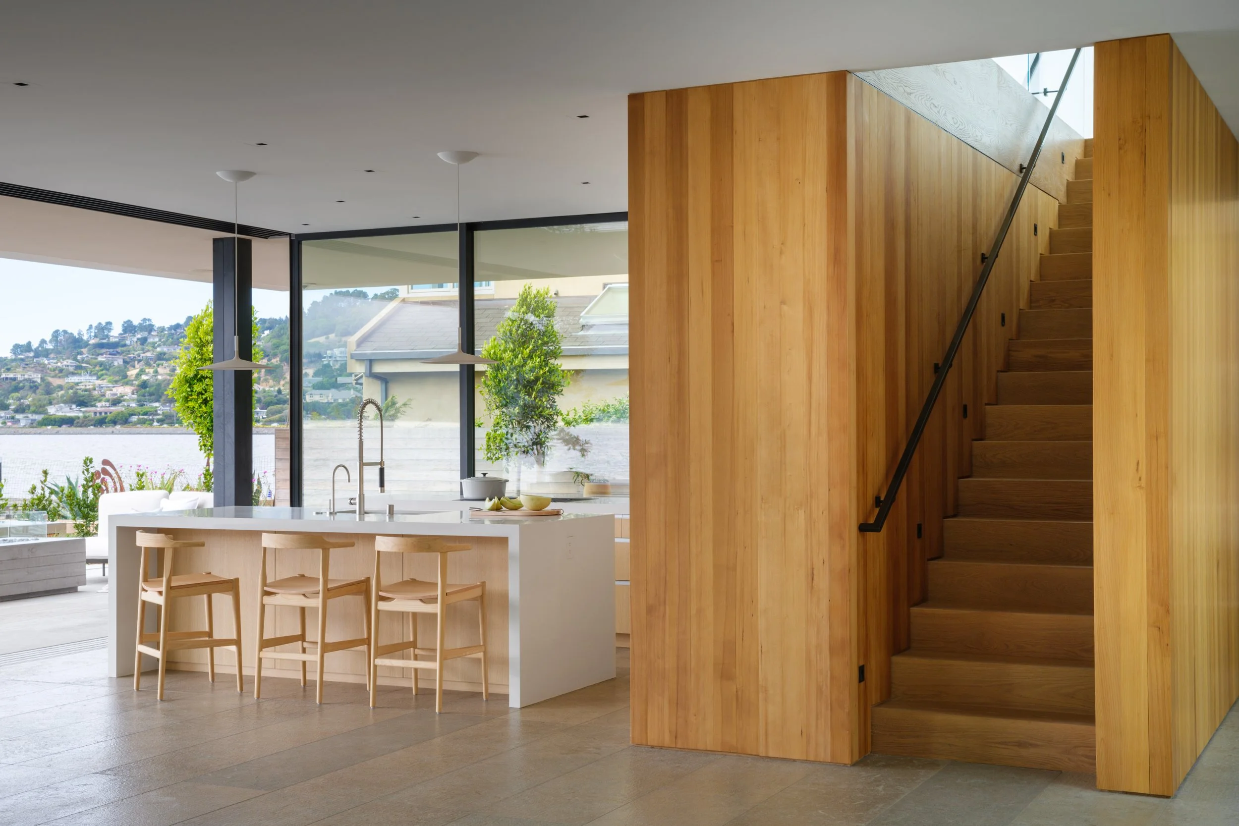 Tiburon waterfront kitchen with white island and three wood stools, framed by large bay view windows and a wood clad staircase core, reflecting modern luxury interior design and indoor outdoor living in Marin County.