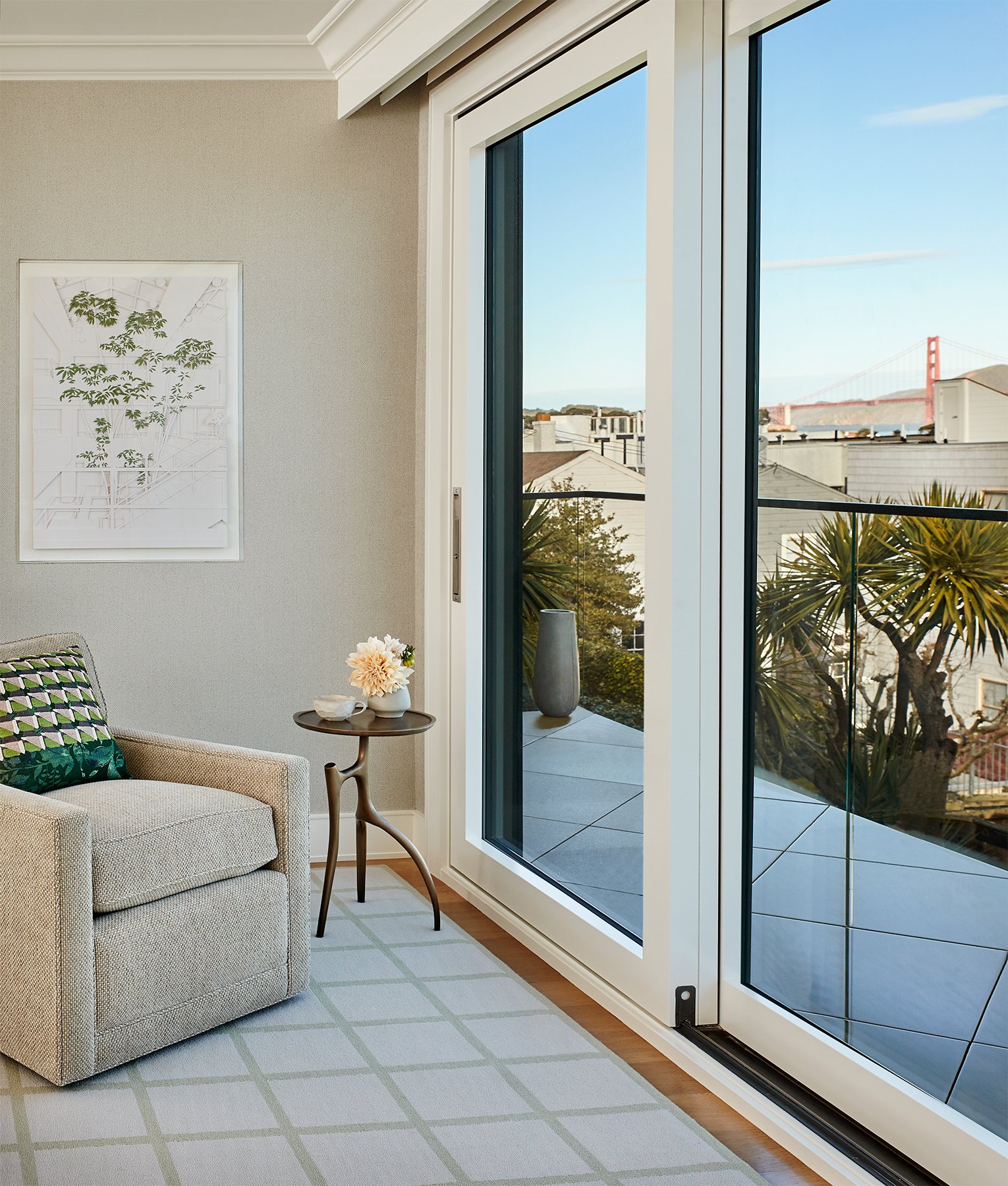Living room with sliding glass door leading to balcony, views of trees, San Francisco bridge in the distance, and modern decor including a beige couch, patterned pillow, small side table with flowers, and wall art on Filbert Street Pacific Heights