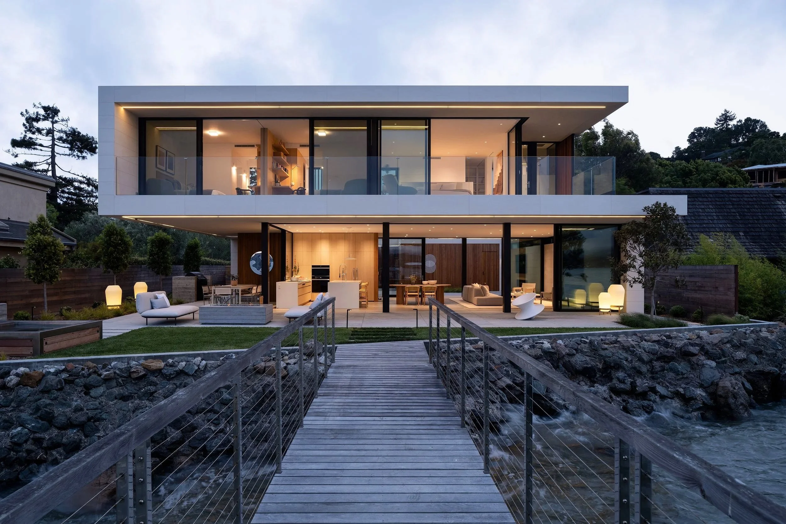 Modern Tiburon waterfront home in Marin County with two story glass walls and bay views, featuring indoor outdoor living across upper and lower decks with curated furnishings, warm ambient lighting, and a wood bridge entry over coastal rocks.