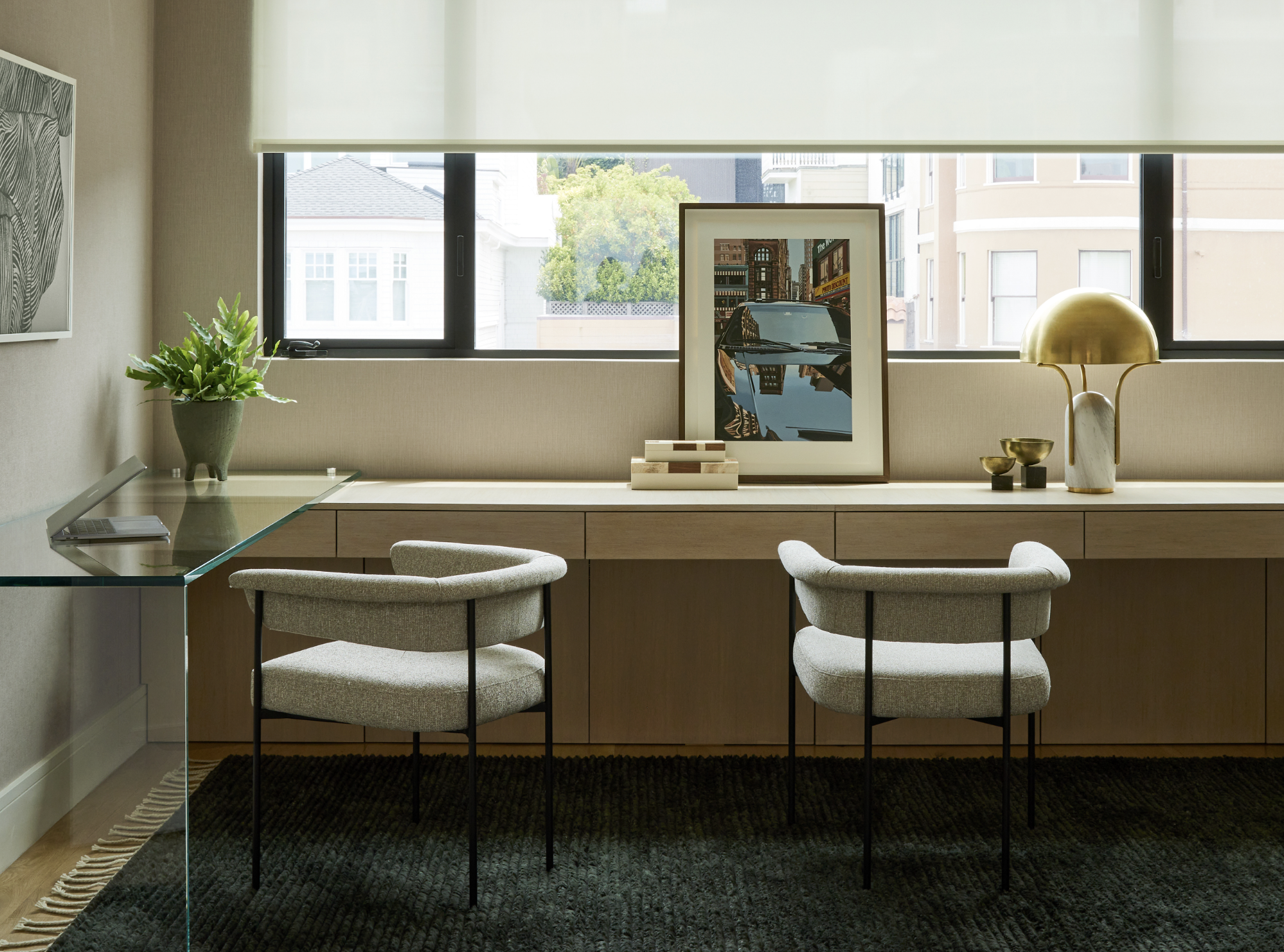 Modern home office in the Marina, San Francisco with a glass desk, artwork curated by BallonStudio two upholstered chairs, decorative items, a framed cityscape painting, a green plant, and a large window with a roller shade