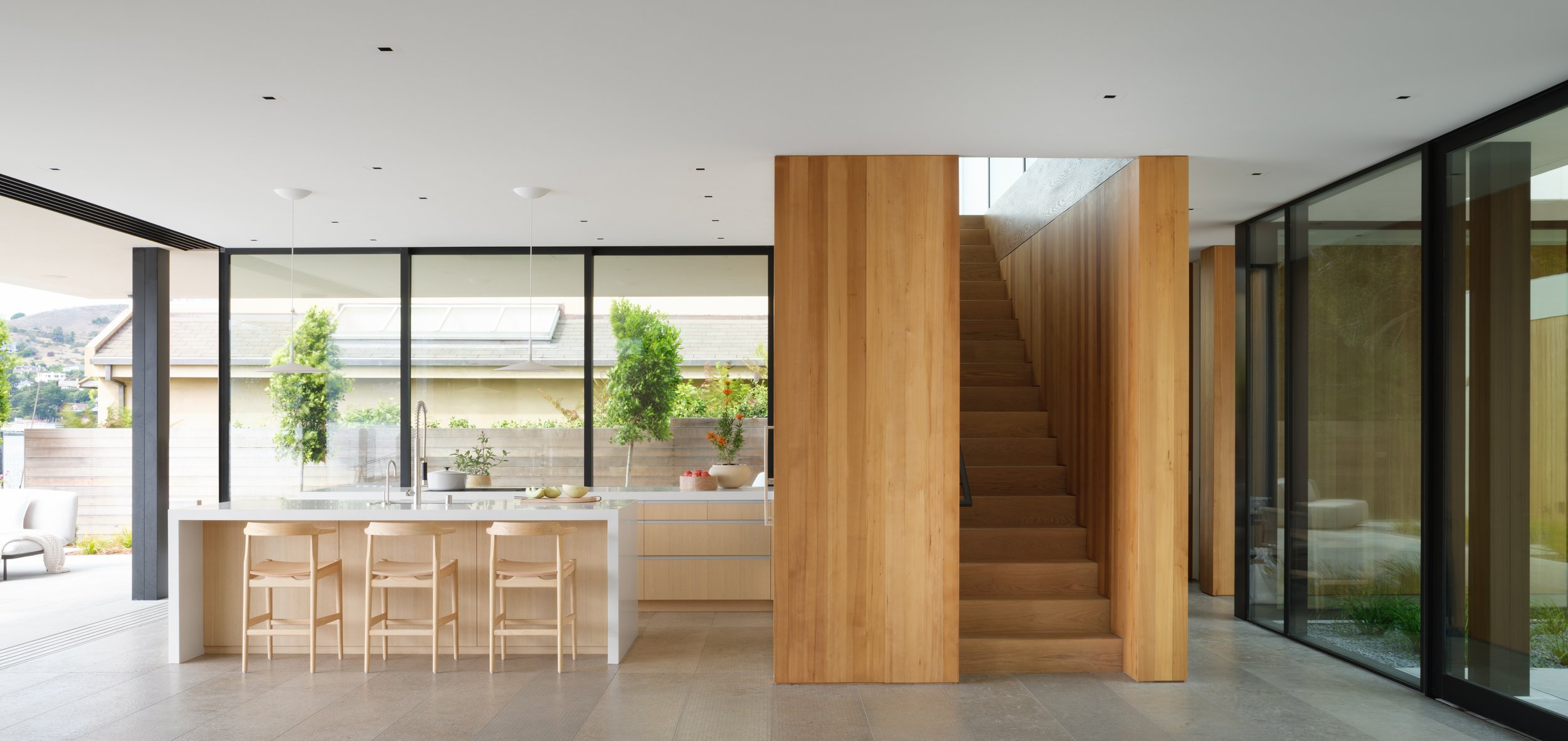 Tiburon waterfront kitchen with large island and warm wood finishes, anchored by a sculptural wood stair volume and floor to ceiling glass that brings in greenery and supports modern Marin County indoor outdoor living.