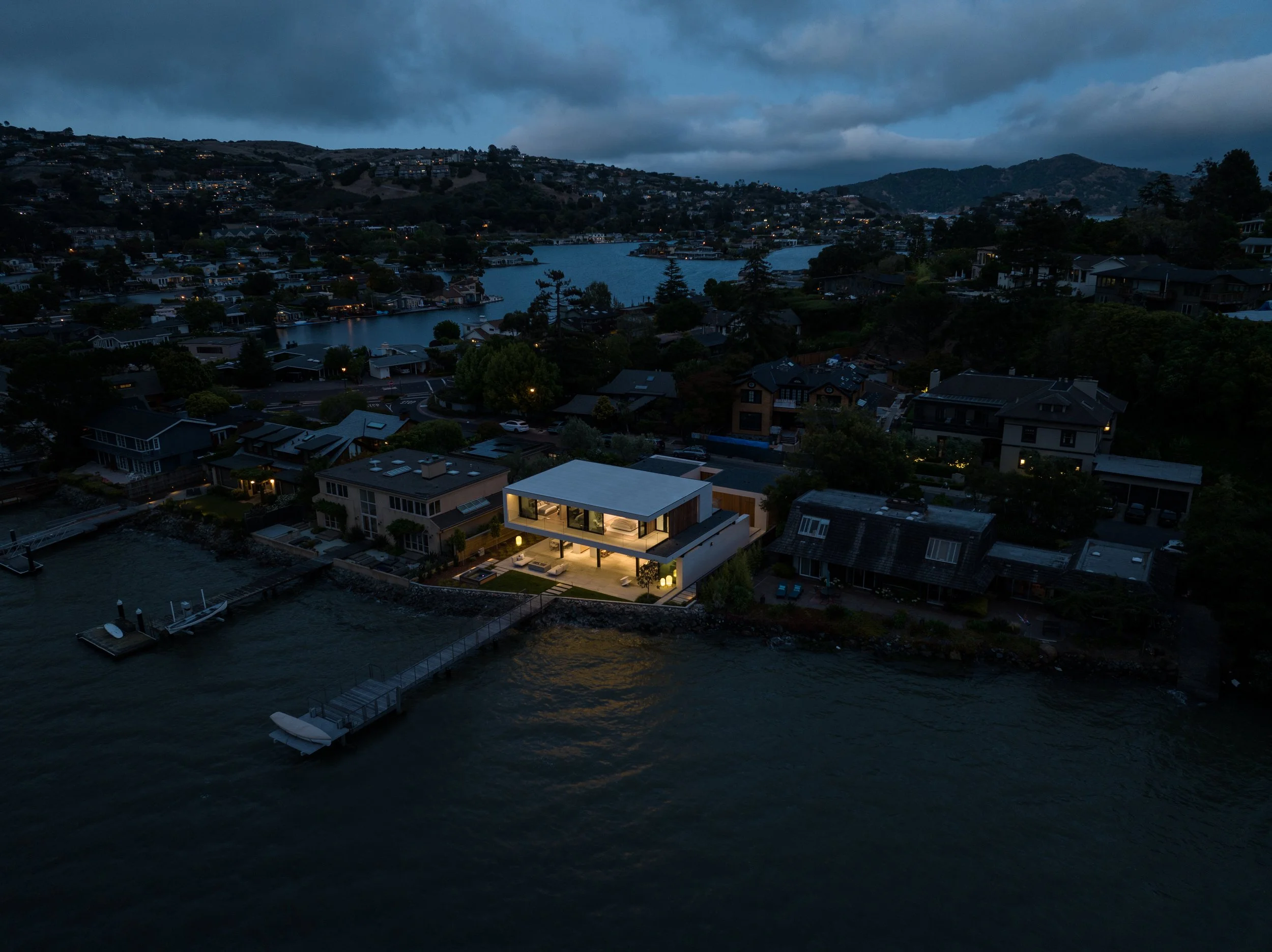 Aerial night view of a modern Tiburon waterfront home in Marin County, glowing beside a dock and boat on the bay, with hillside neighborhood lights under cloudy skies and sweeping San Francisco Bay views.