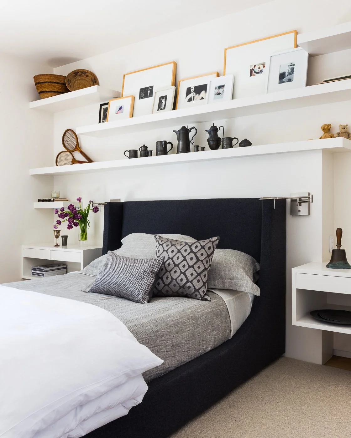 St. Helena Napa Valley bedroom with a dark upholstered headboard and layered pillows, styled with white nightstands, flowers, and books, plus built in shelves displaying family photos and collected objects for warm luxury living.
