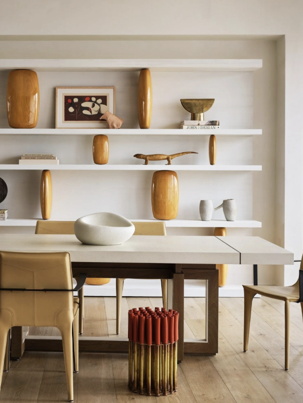 Modern dining room with a light-colored table, beige and black chairs, and decorative shelving with vases, a framed artwork, a sculpture, and books.