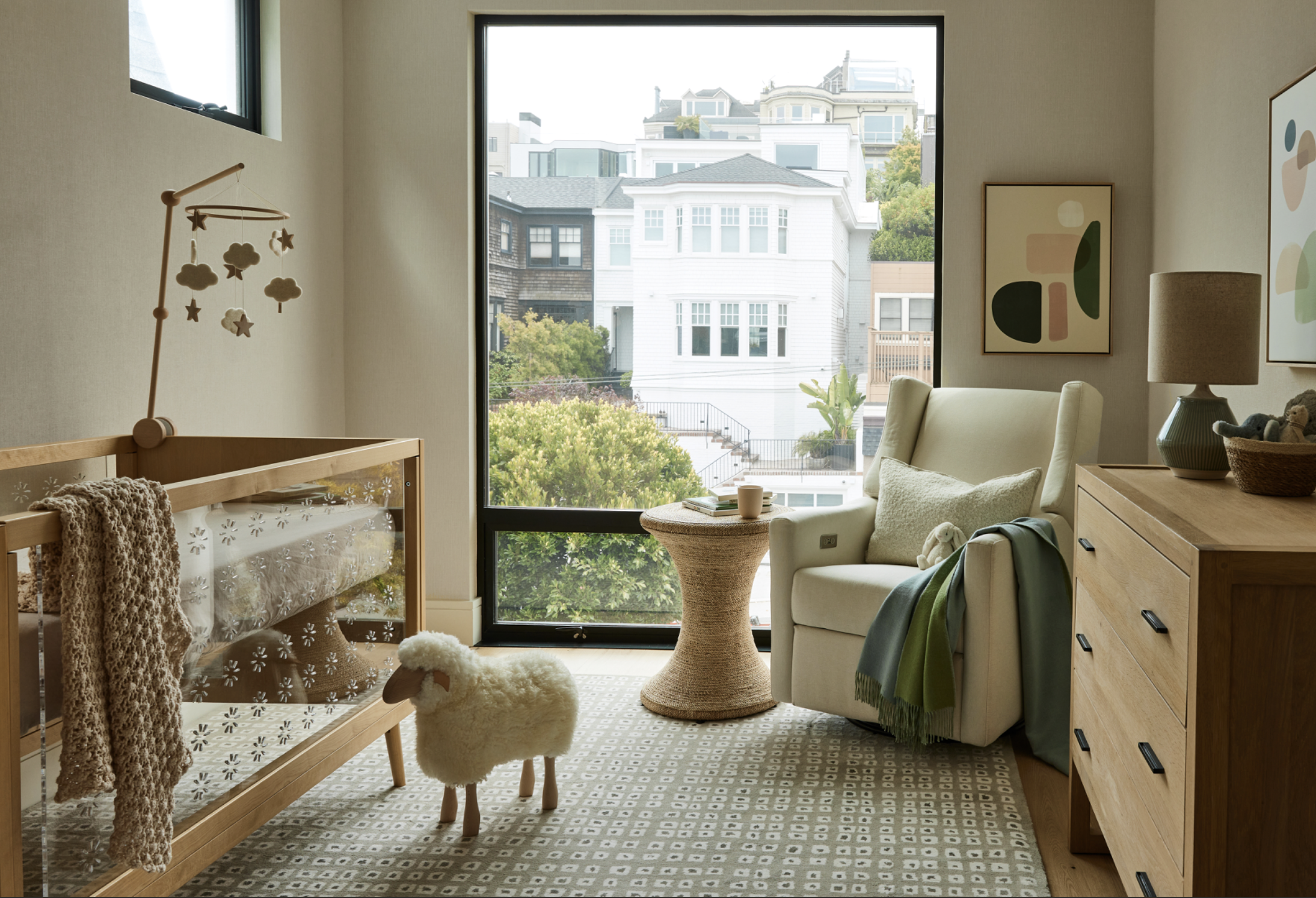 A nursery room with a wooden crib, a soft sheep toy with wooden legs in front of the crib, a white armchair with a pillow and a blanket, a small round side table with a mug, a dresser with a lamp and basket of toys, two pieces of abstract artwork on the wall, and a large window showing a view of neighboring houses and trees.