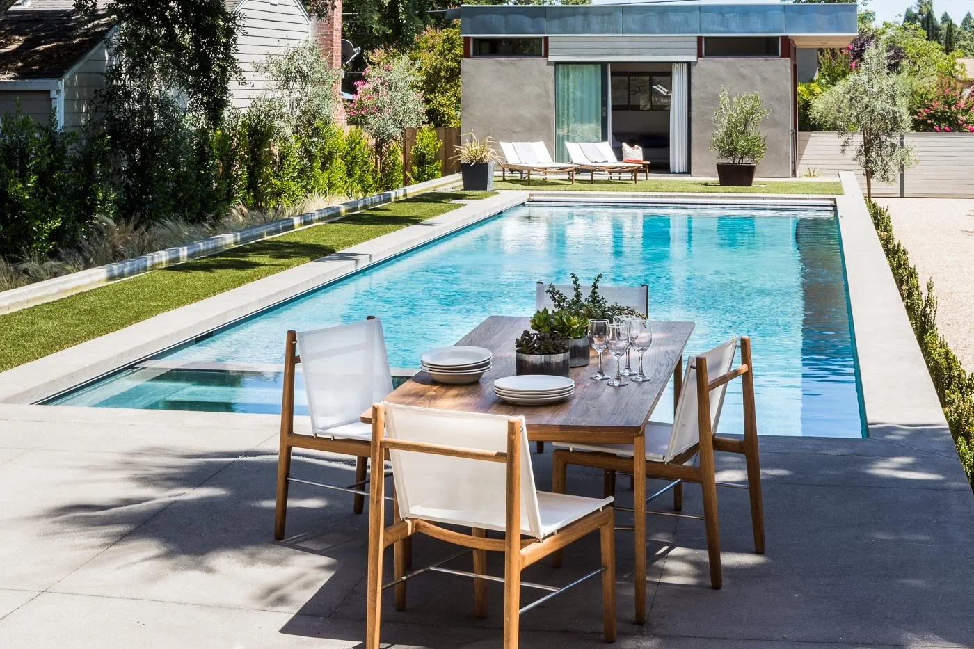 St. Helena Napa Valley backyard with a pool, outdoor dining table set with plates and glasses, potted plants, and lounge chairs near a modern guesthouse, framed by trees for luxury indoor outdoor living.