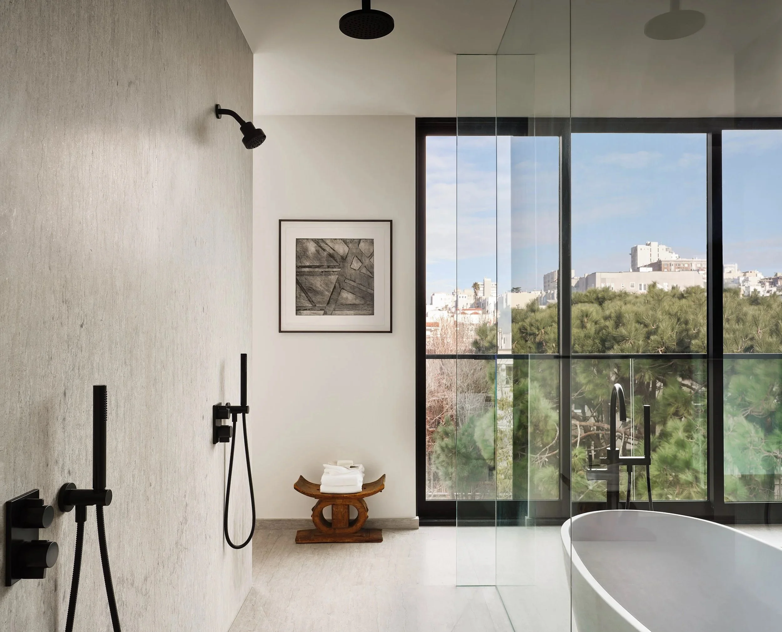 Broadway Penthouse Pacific Heights bathroom with a freestanding tub and matte black fixtures, framed by glass and natural stone textures, with art on the wall and a window view of trees and San Francisco buildings.