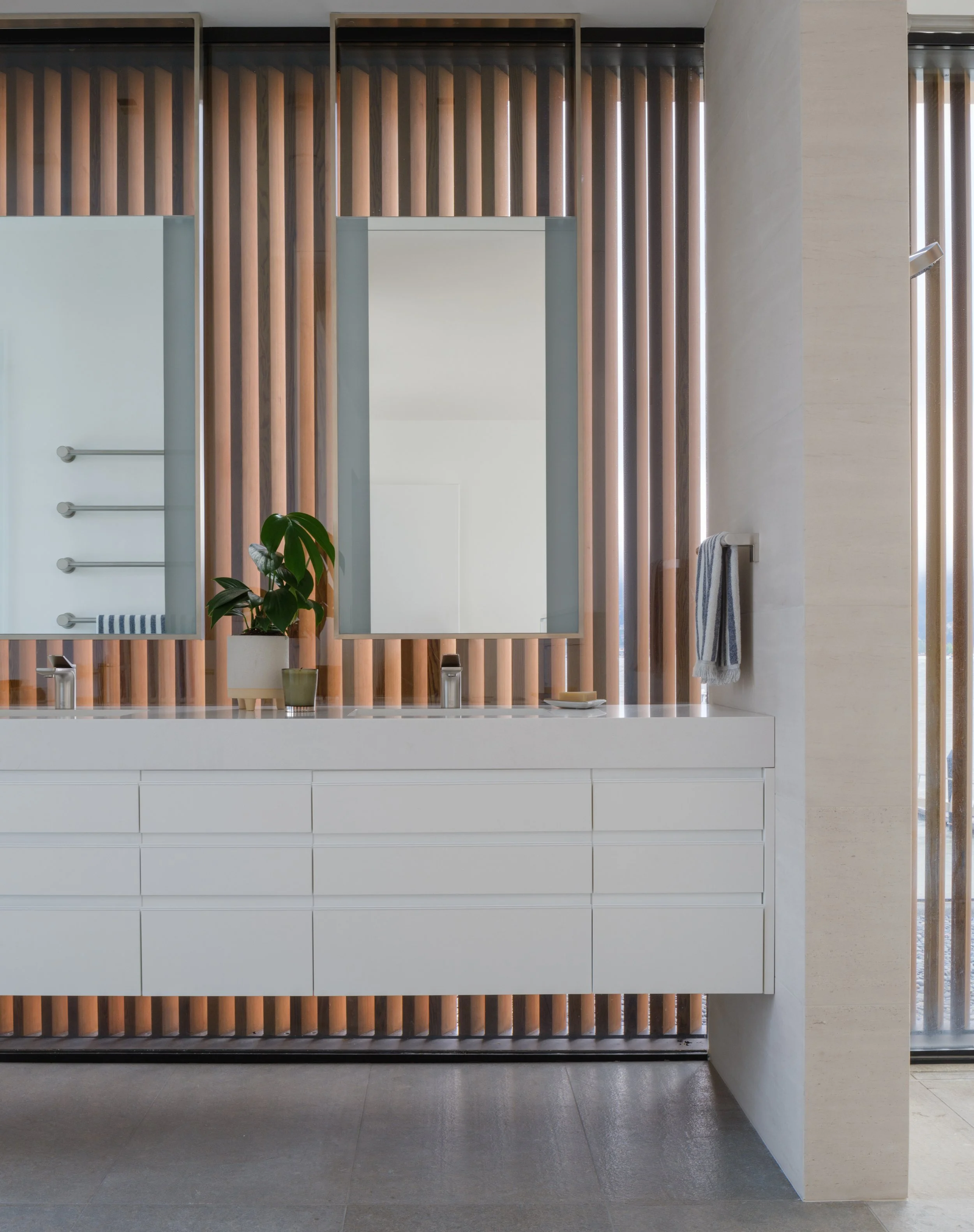 Tiburon waterfront bathroom with white double vanity and dual mirrors, styled with a potted plant and towel hook, featuring warm wood slat accents and a bright window for modern Marin County luxury interior design.